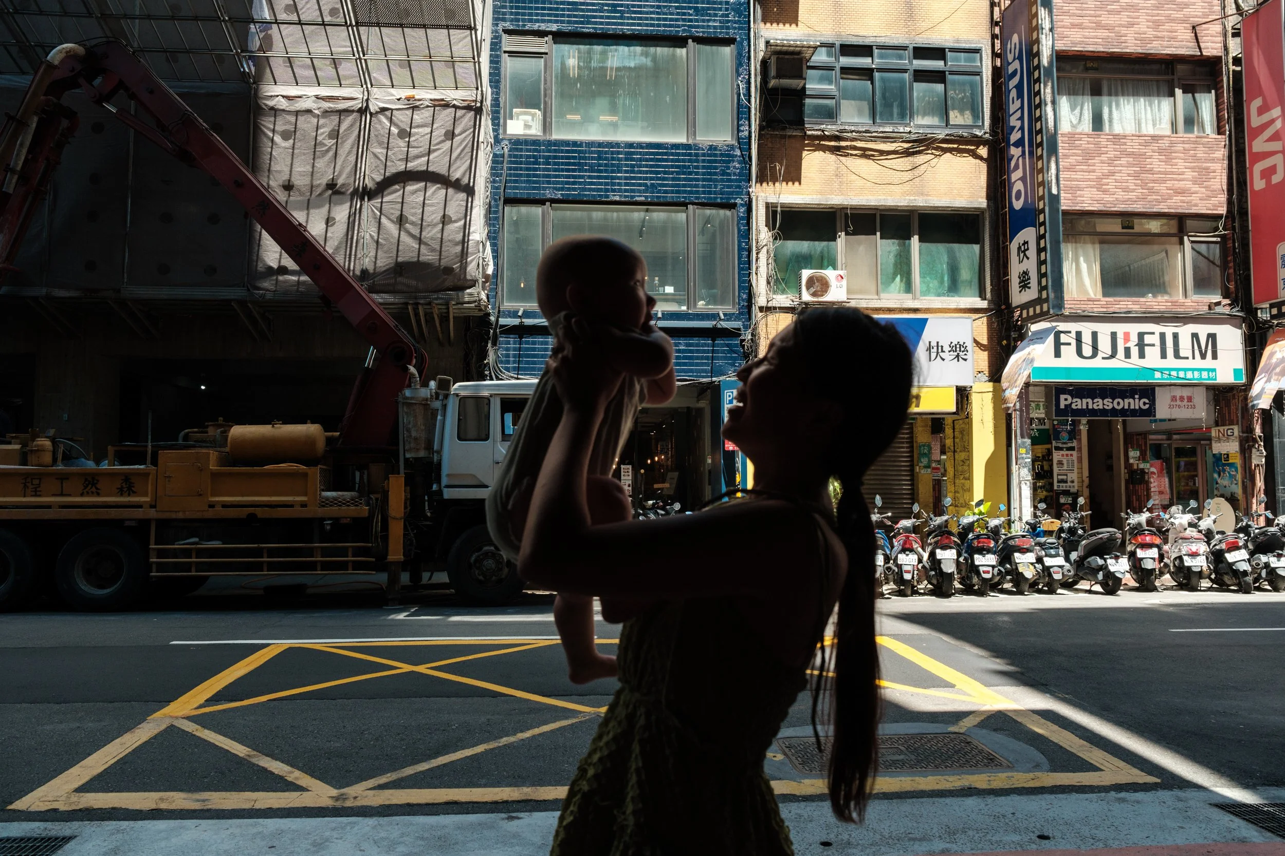 A woman holds her baby up and looks in his eyes. A colorful line of shops is in the background. Photographed by Taipei photographer Cahleen Hudson.