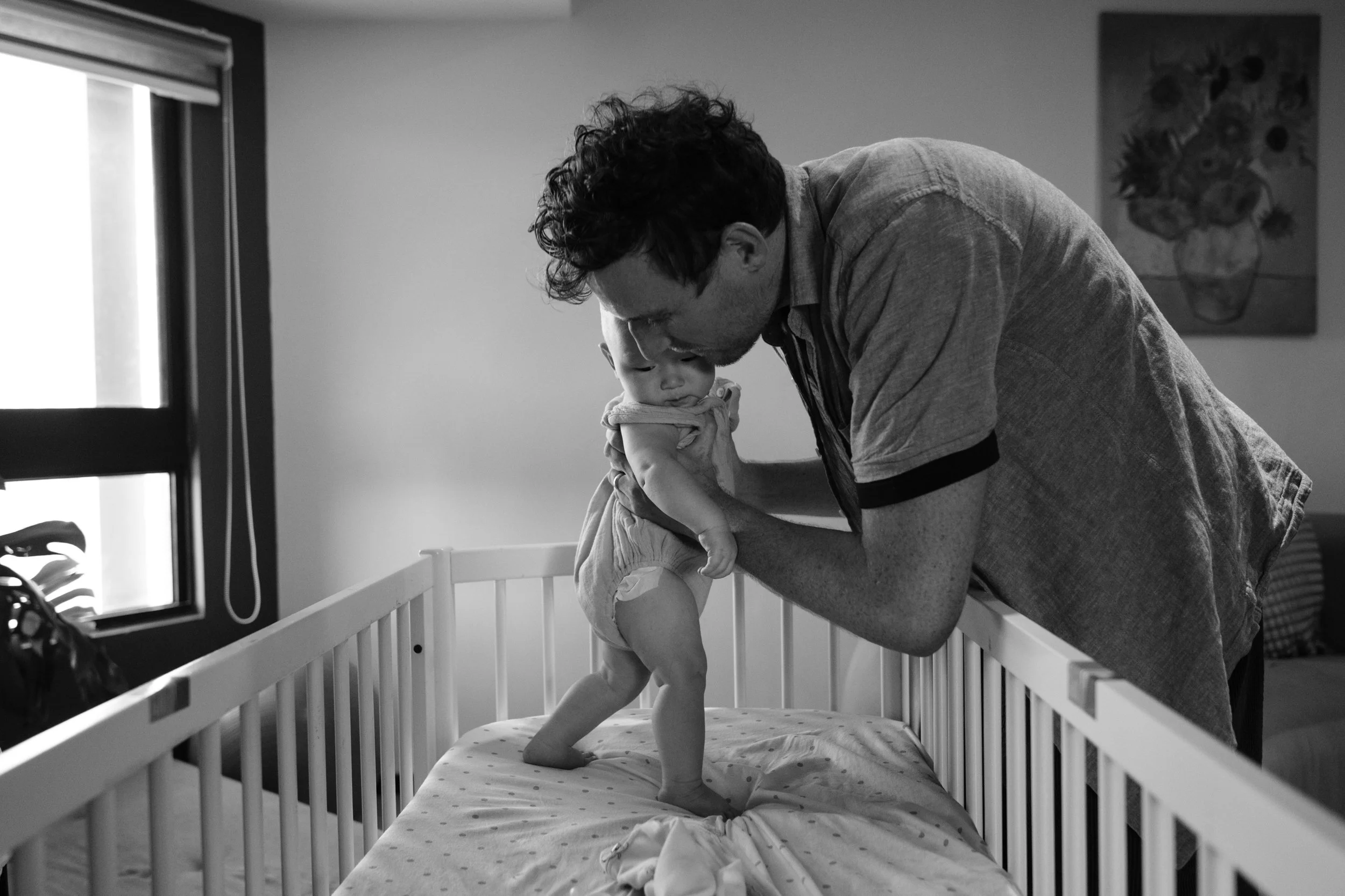A black and white photo of a man putting his baby in a crib. Photographed by Taipei documentary family photographer Cahleen Hudson.