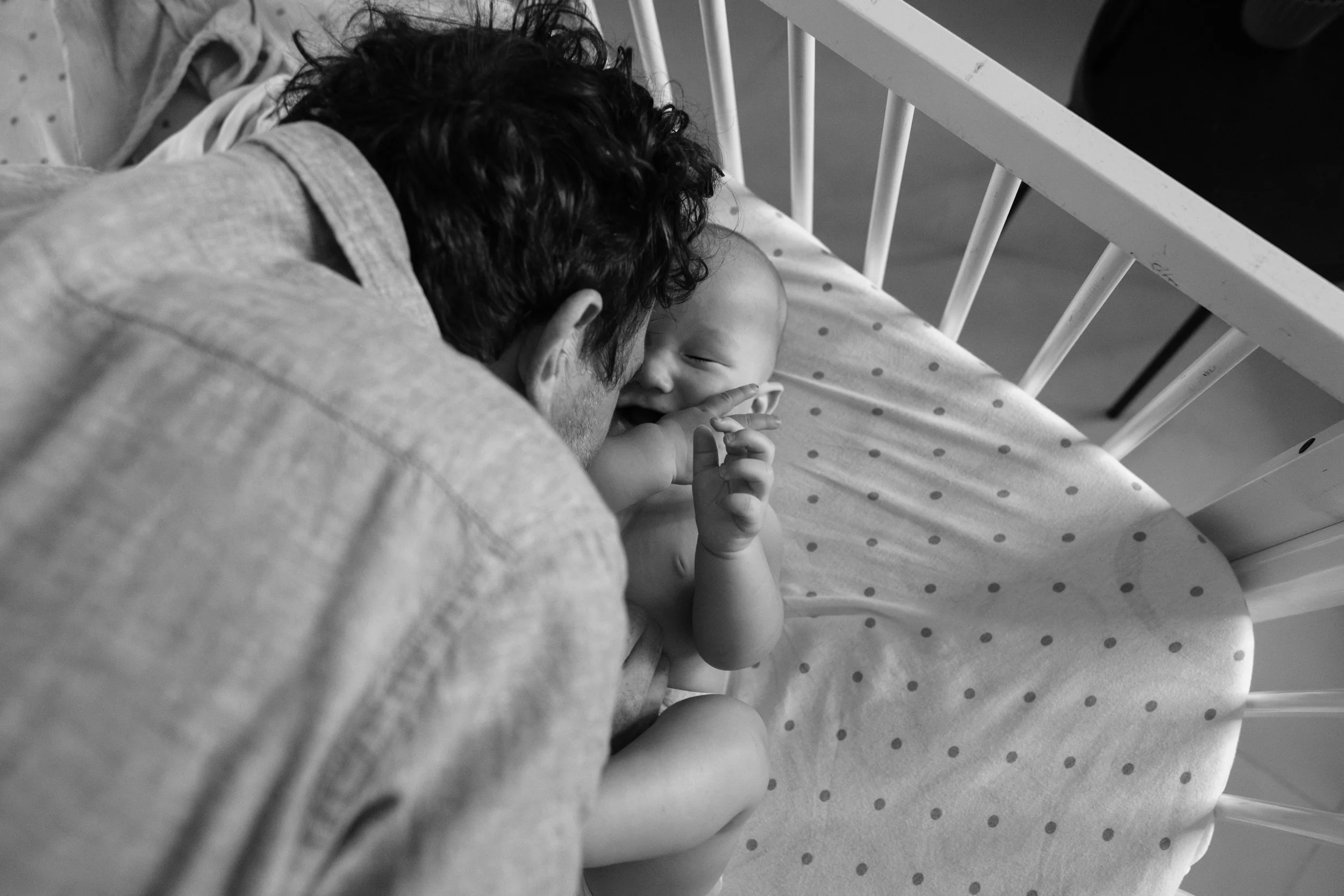 A black and white photo of a man leaning down and kissing his baby in a crib. The baby is closing his eyes and laughing.