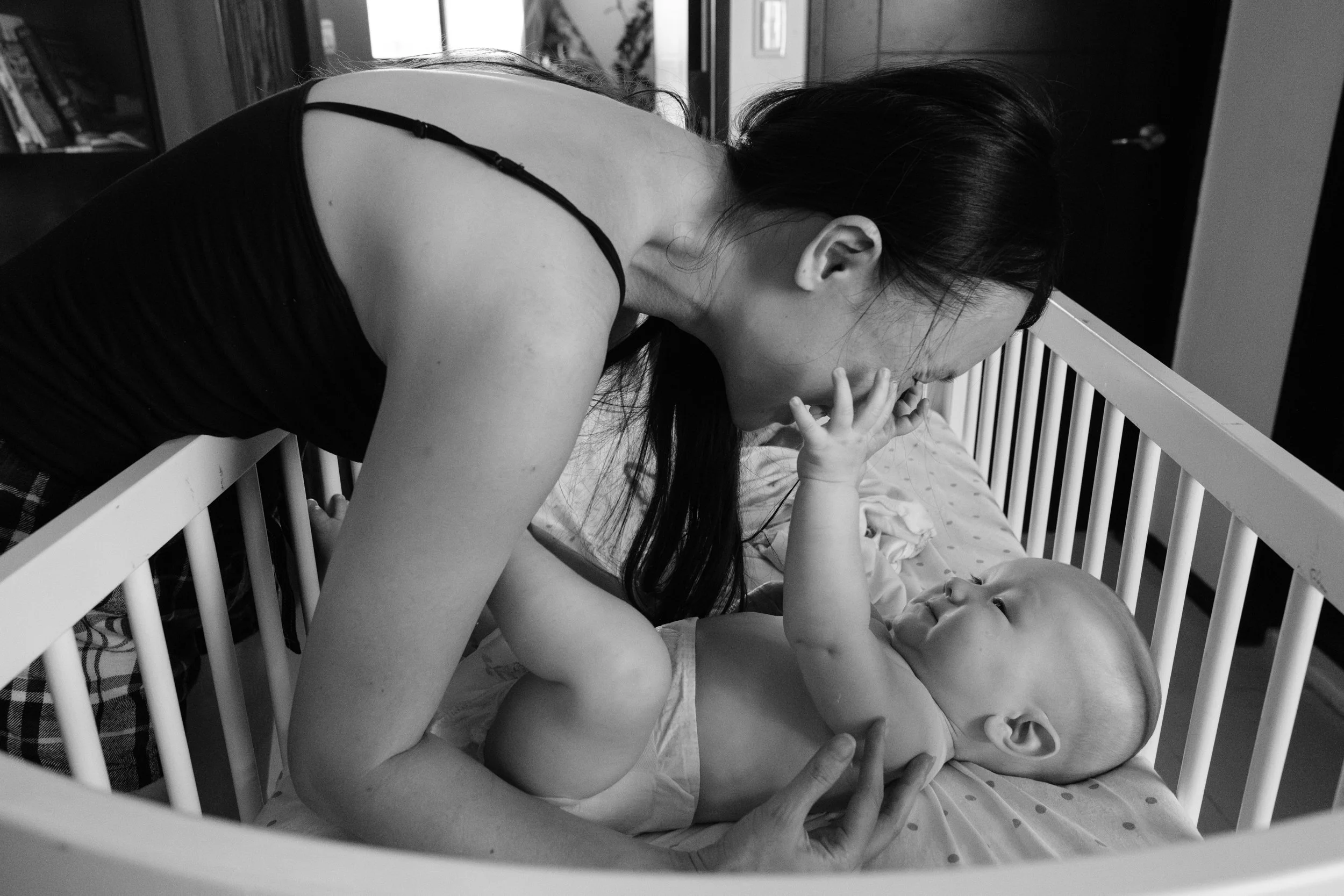 A black and white photo of a woman leaning over and nuzzling her baby in a crib. The woman is laughing because the baby is reaching up and grabbing her face.