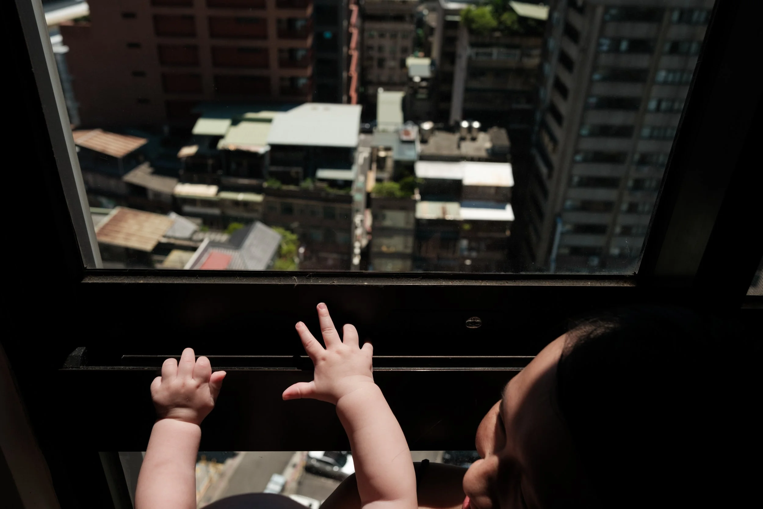 A color photo of a baby's hands reaching towards the window. Light is shining on the baby's hands and the Taipei cityscape can be seen out the window. 