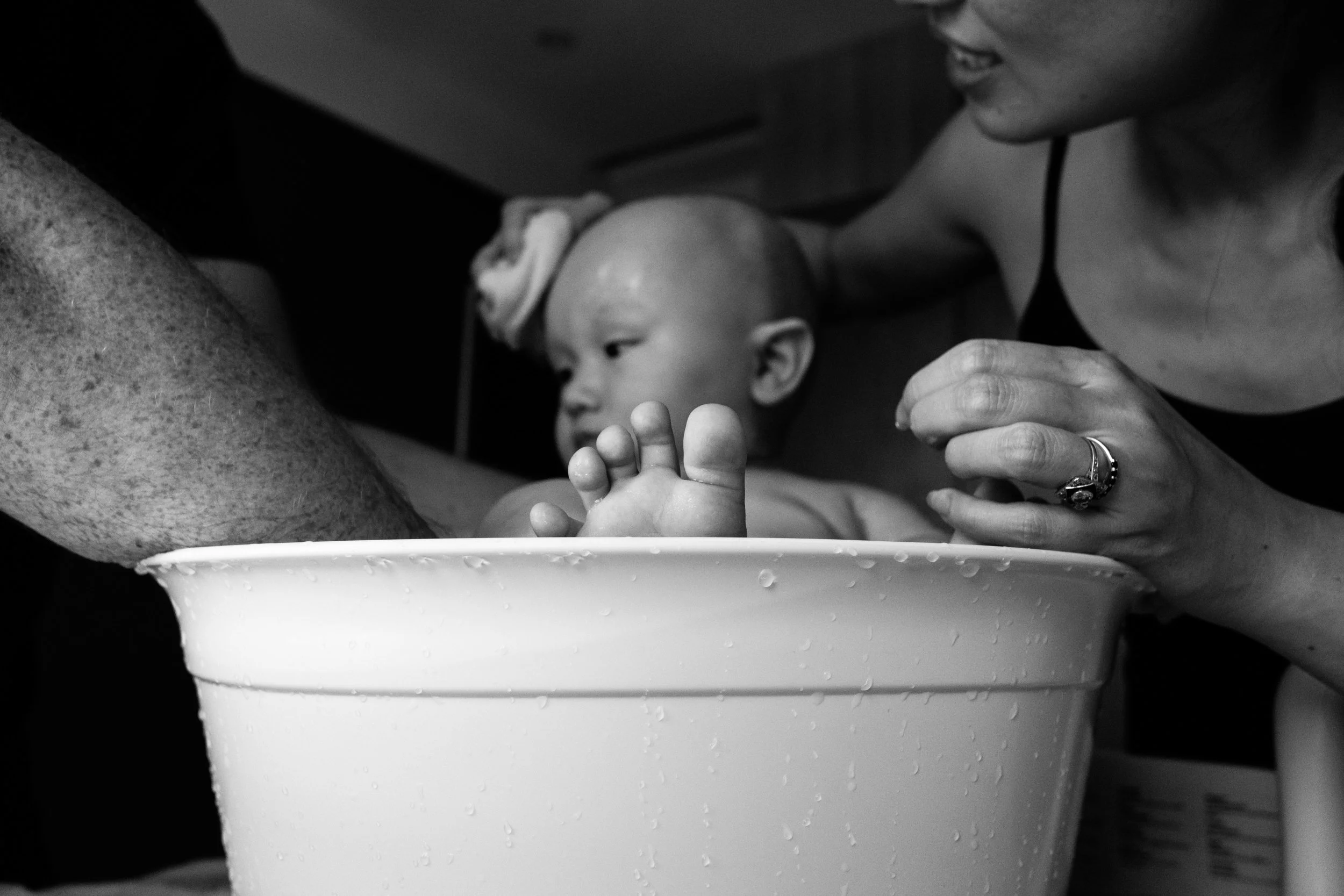A black and white photo of a baby sitting in a small bathtub. The focus is on his toes sticking above the rim of the bathtub in the foreground.