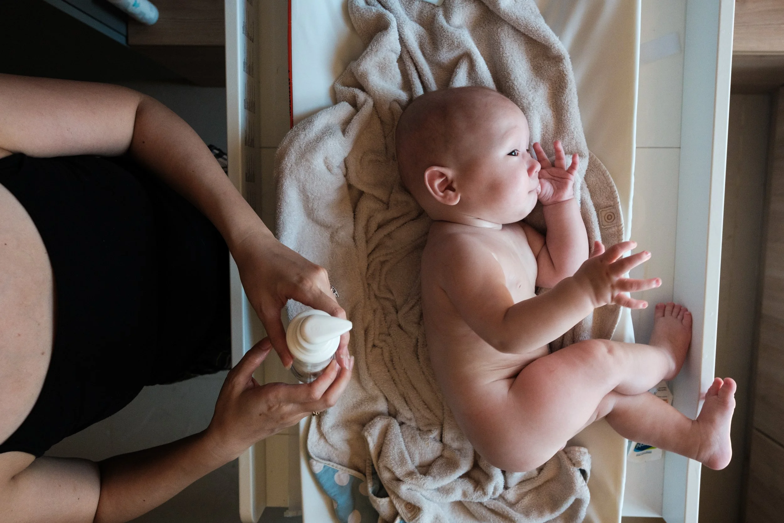 A color photo of a baby having lotion put on him after a bath. The baby is on his side and looking out the window. Light is shining on the baby from the window.