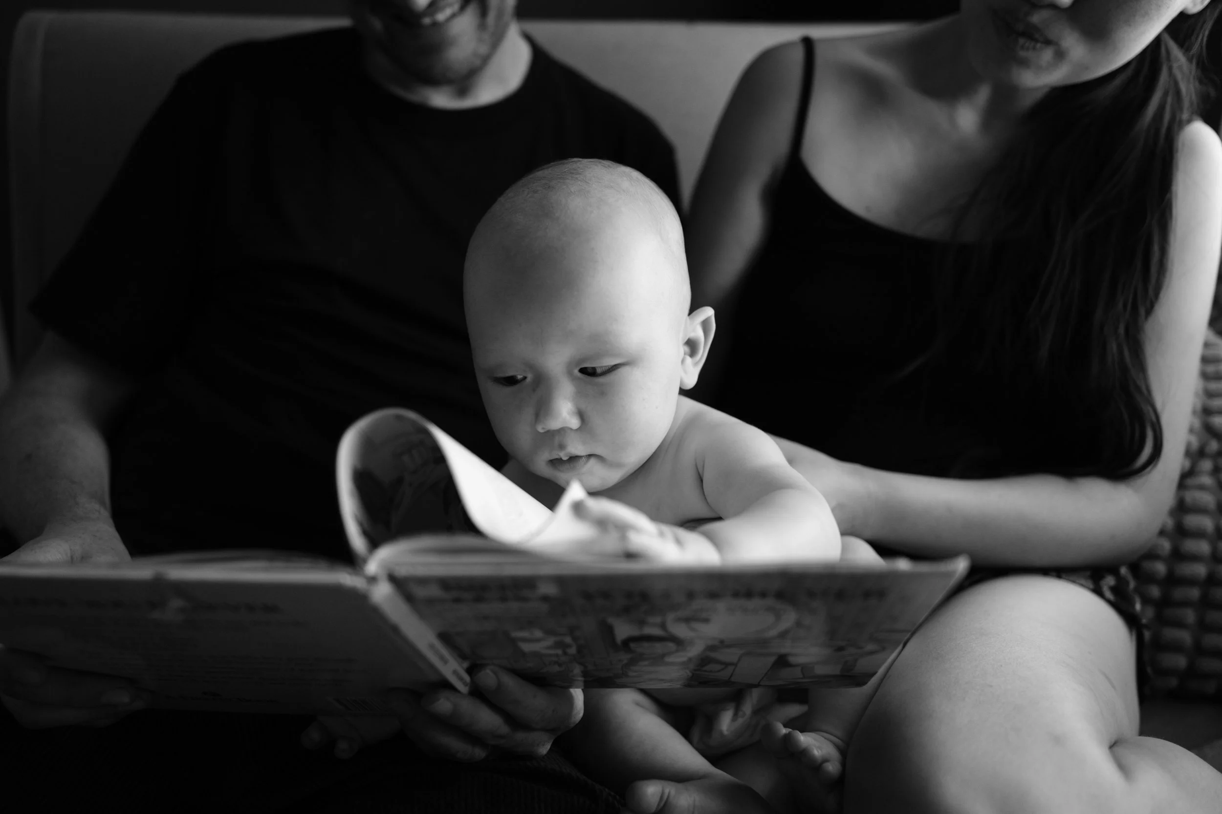 A black and white photo of a baby turning the pages of a picture book. His parents are reading the book to him and can be seen in the background.