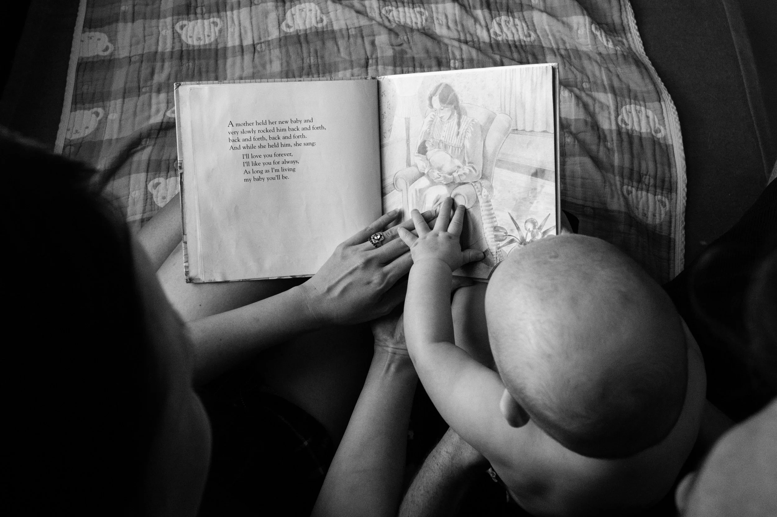 A black and white photo of a baby's hand resting on his mother's hand as she turns the pages of a picture book she's reading him. 