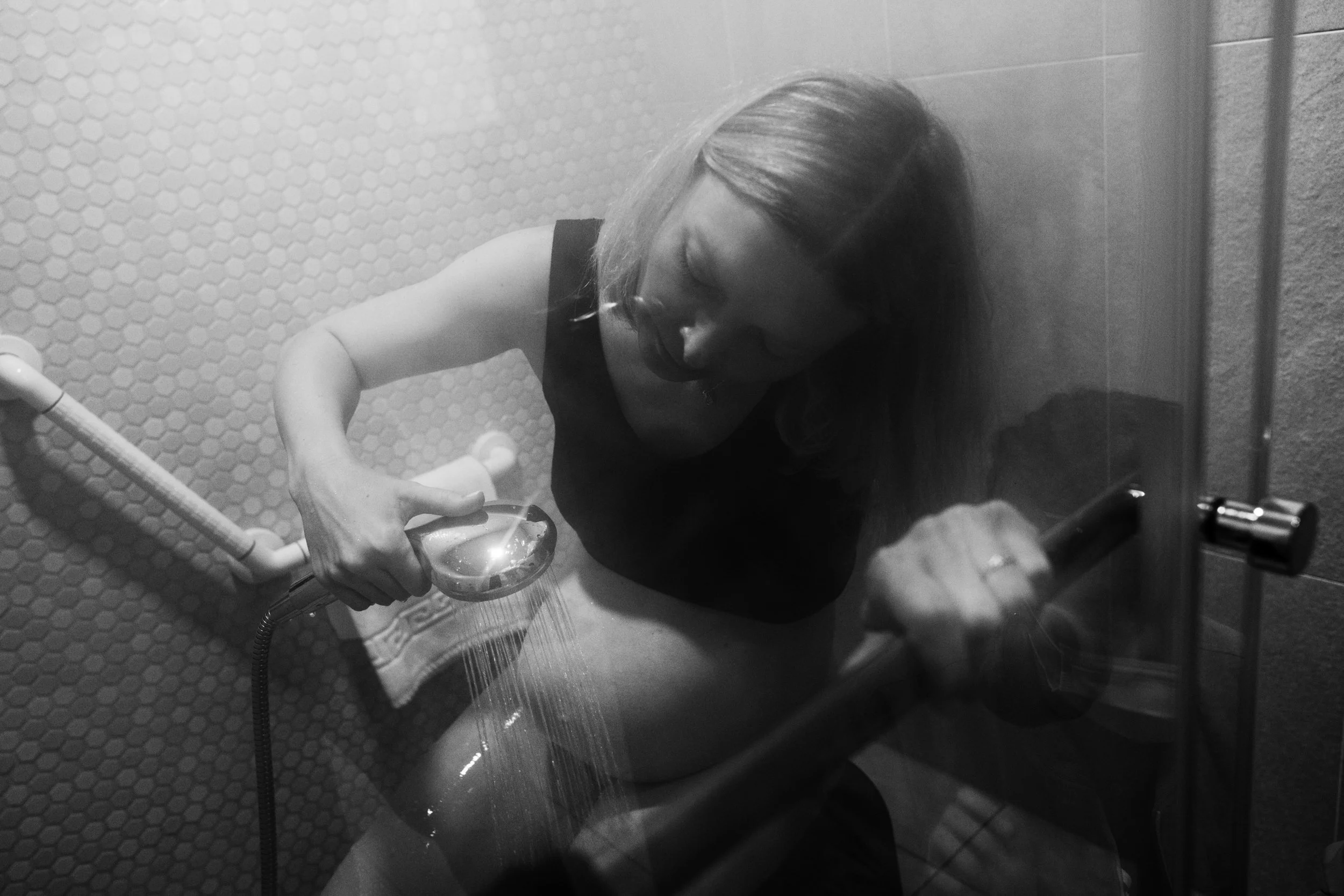 A black and white photo of a woman in labor in the shower at Dianthus in Taipei. She's spraying warm water on her stomach.