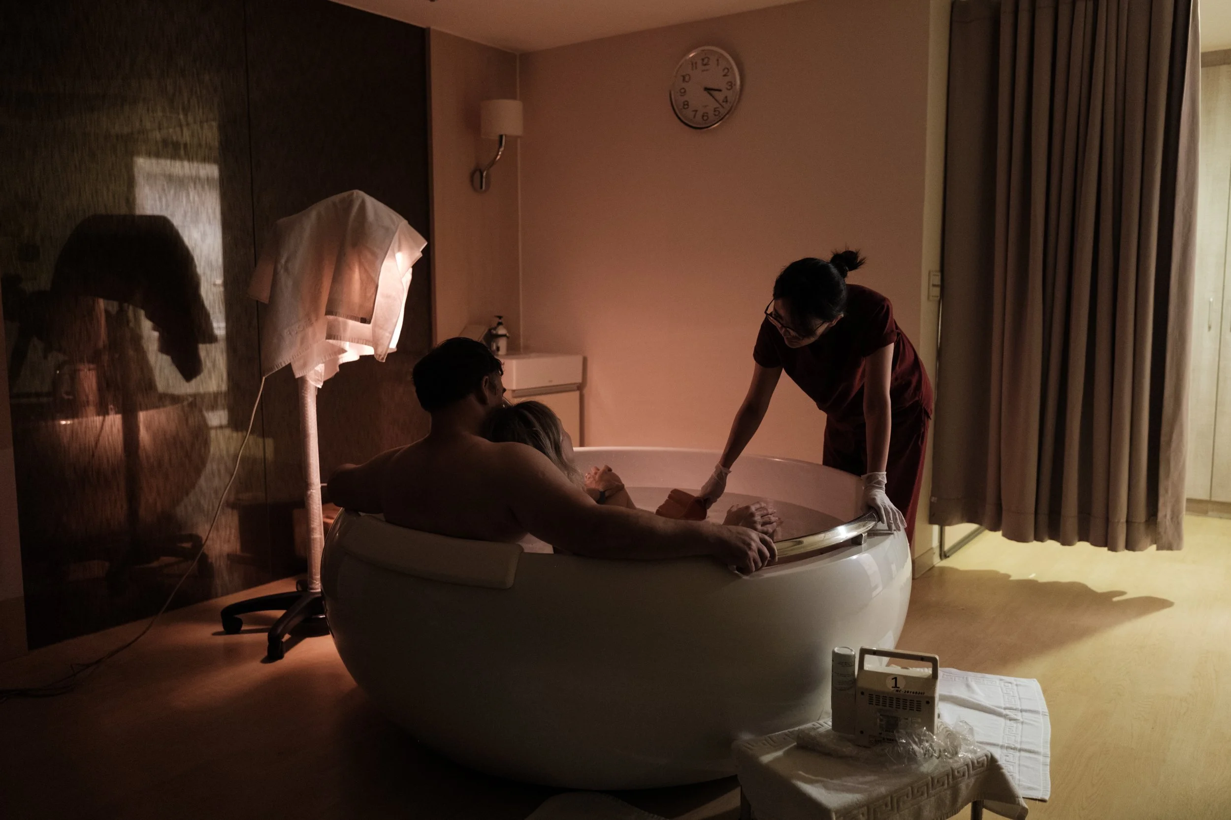 A color photo of a nurse at Dianthus in Taipei leaning over a birthing tub and assisting a couple. A woman is in labor in the tub and leaning back against her husband.