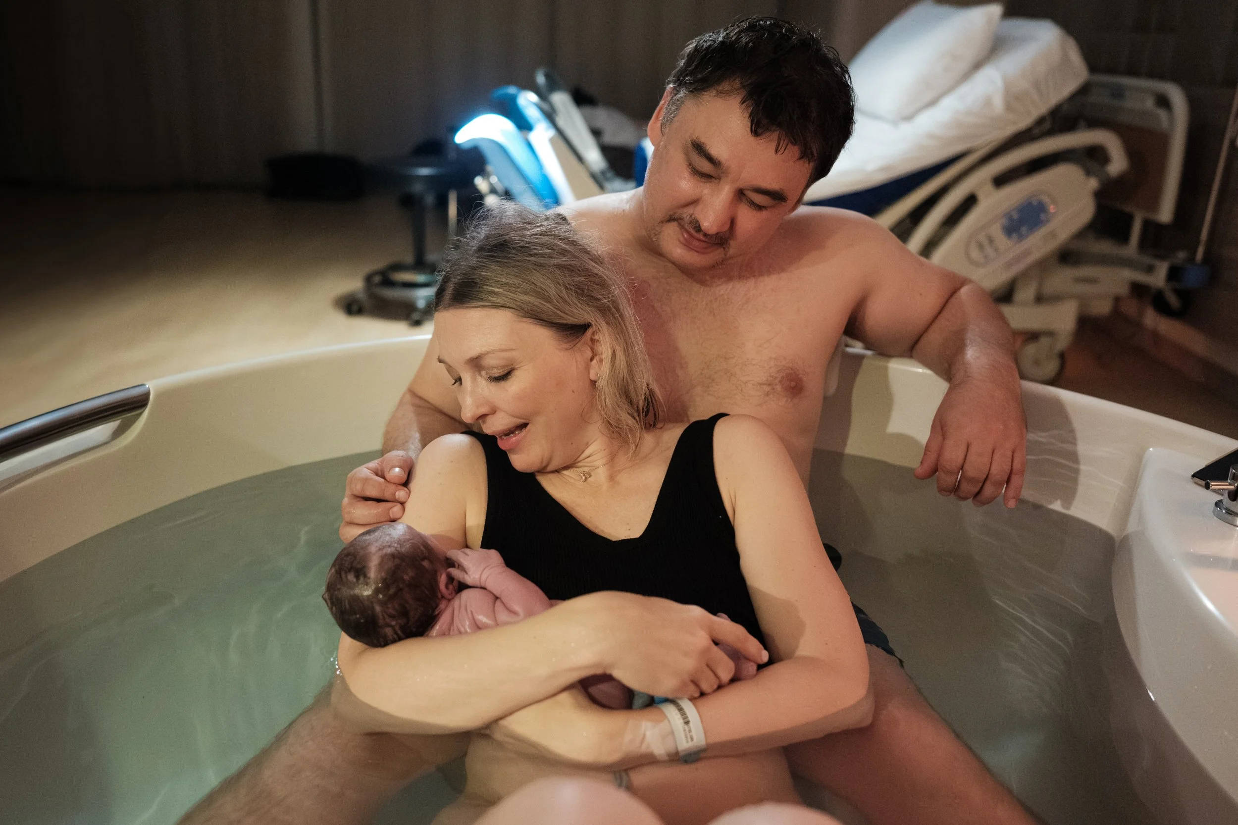 A color photo of a woman leaning against her husband in a birthing tub in Dianthus and holding her newborn baby. She is looking down and smiling at her baby.
