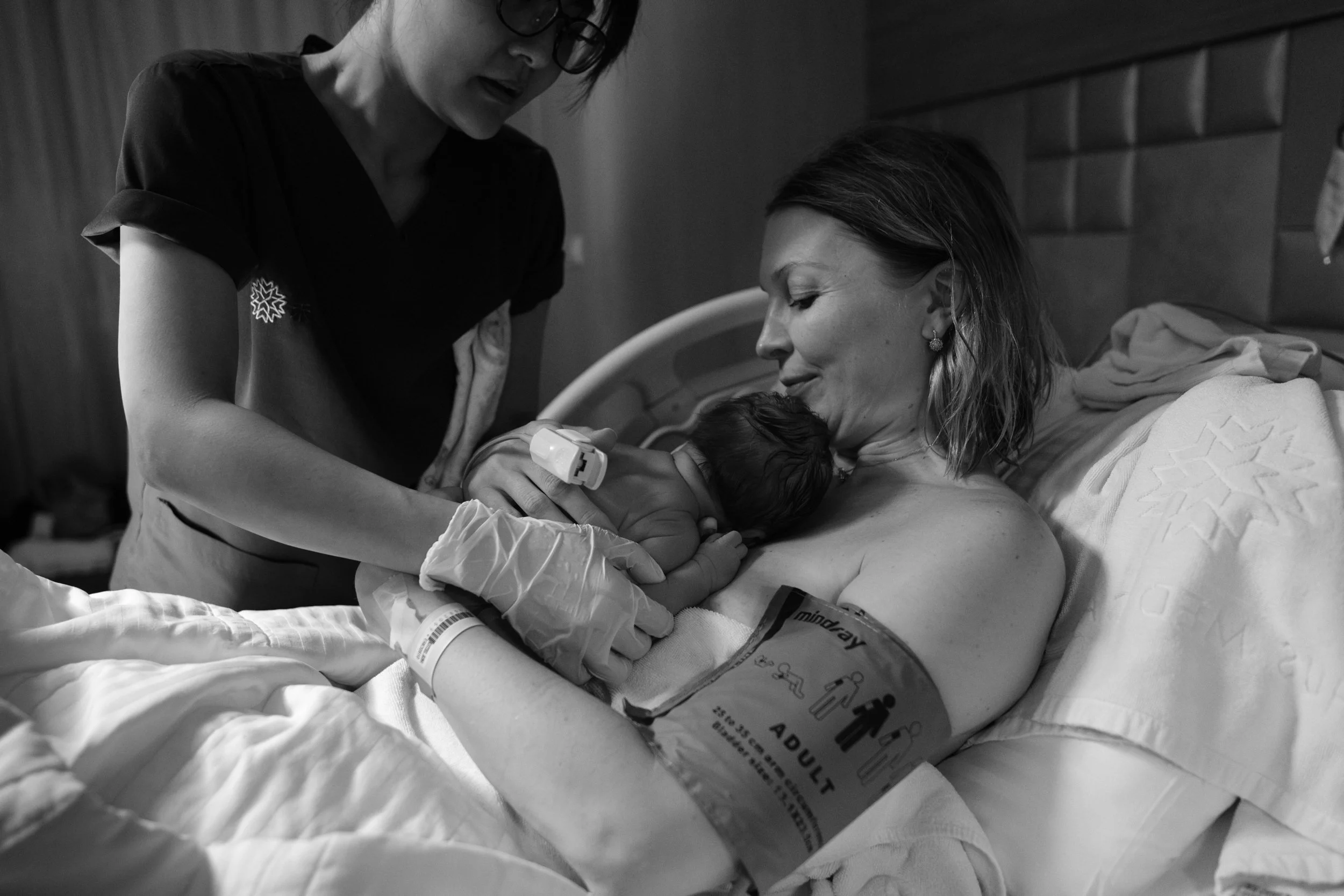 A black and white photo of a nurse handing a newborn baby to her mom, who is sitting up in a hospital bed at Dianthus in Taipei, Taiwan.