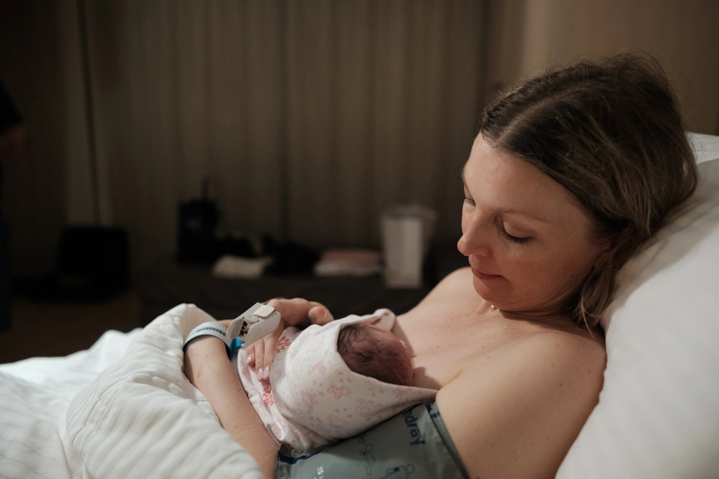 A color photo of a mother holing her newborn baby in a hospital bed at Dianthus in Taipei, taiwan. She is gazing down at her baby and smiling.