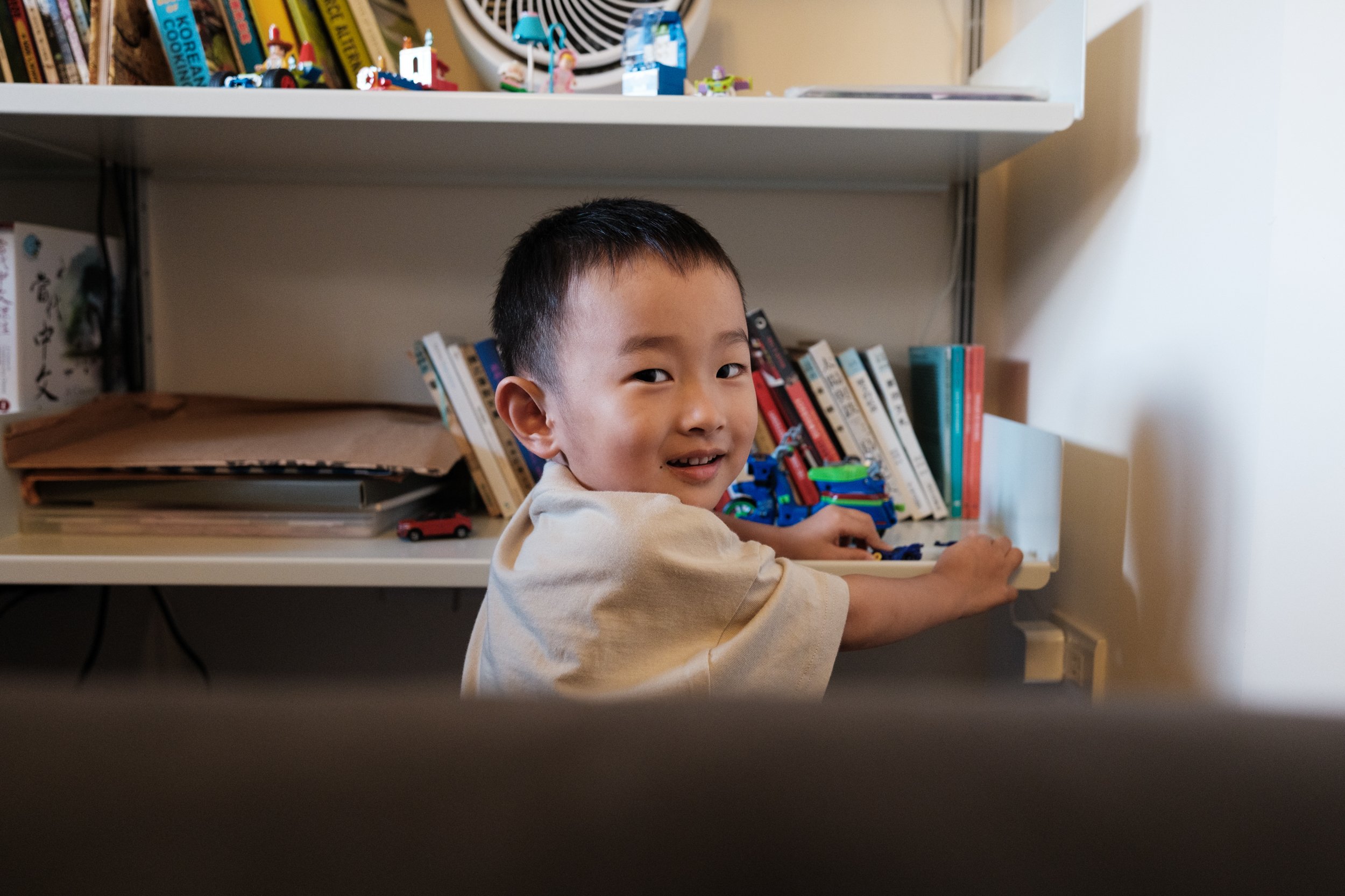 A color image of a little boy getting something off a bookshelf in his living room in Taipei, Taiwan. The boy is looking at the camera and smiling.