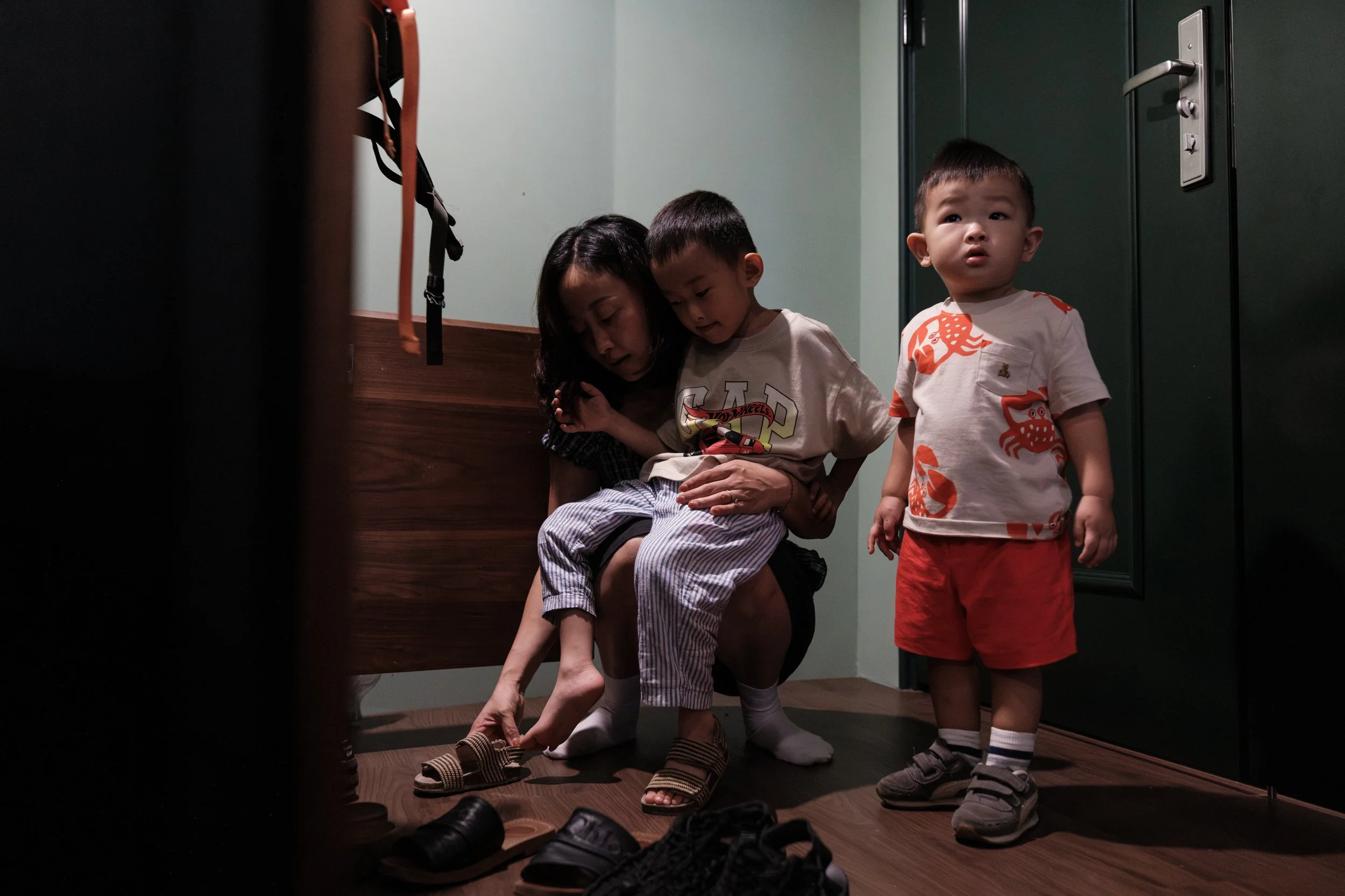 A color image of a mom helping her two young sons put on their shoes before they go outside to play in their Taipei neighborhood. They are near the front door of their apartment.