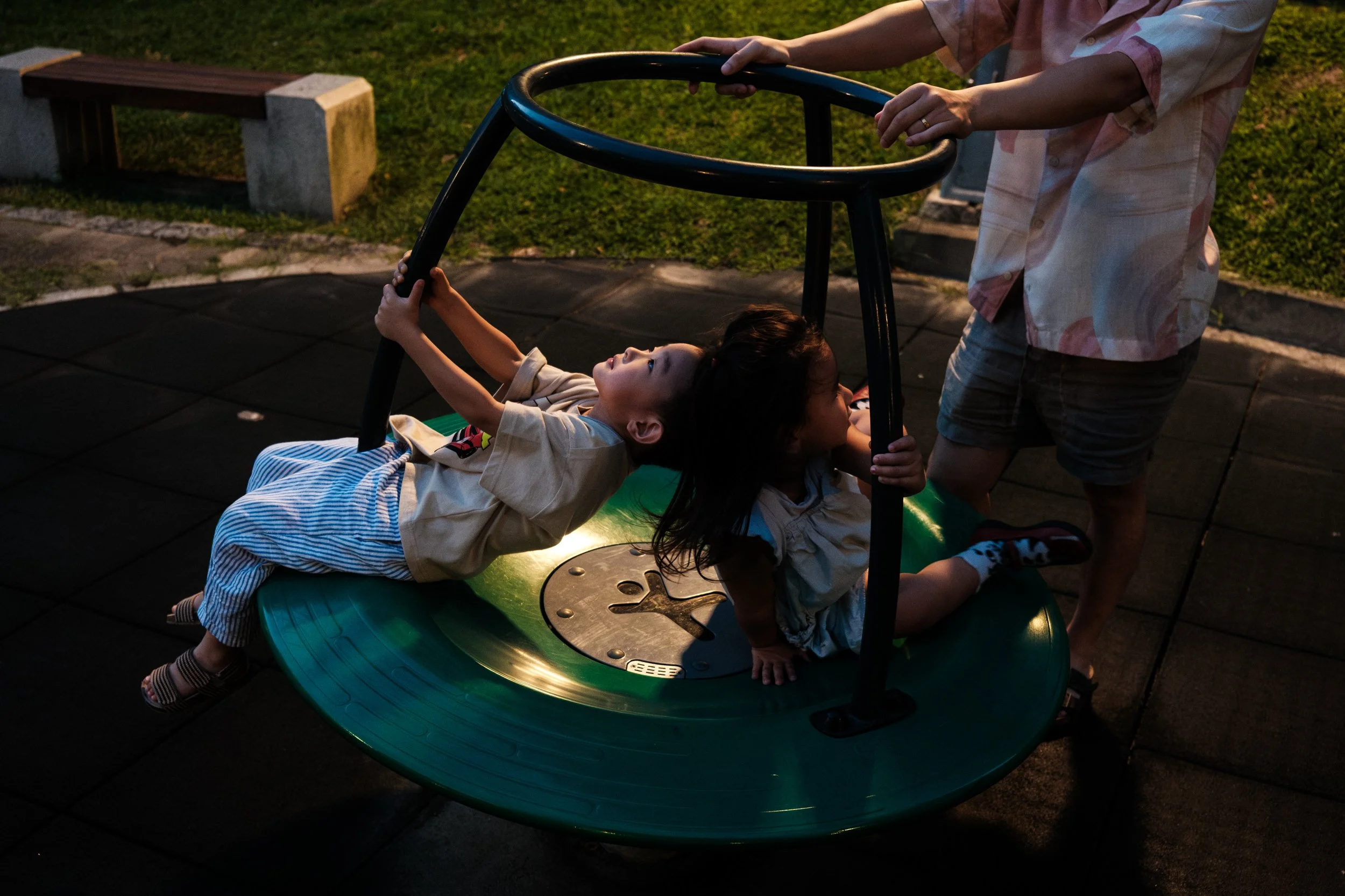 A color photo of a little boy and girl on a merry-go-round in a Taipei park at night. The kids are looking up towards a street lamp, casting light on their faces.