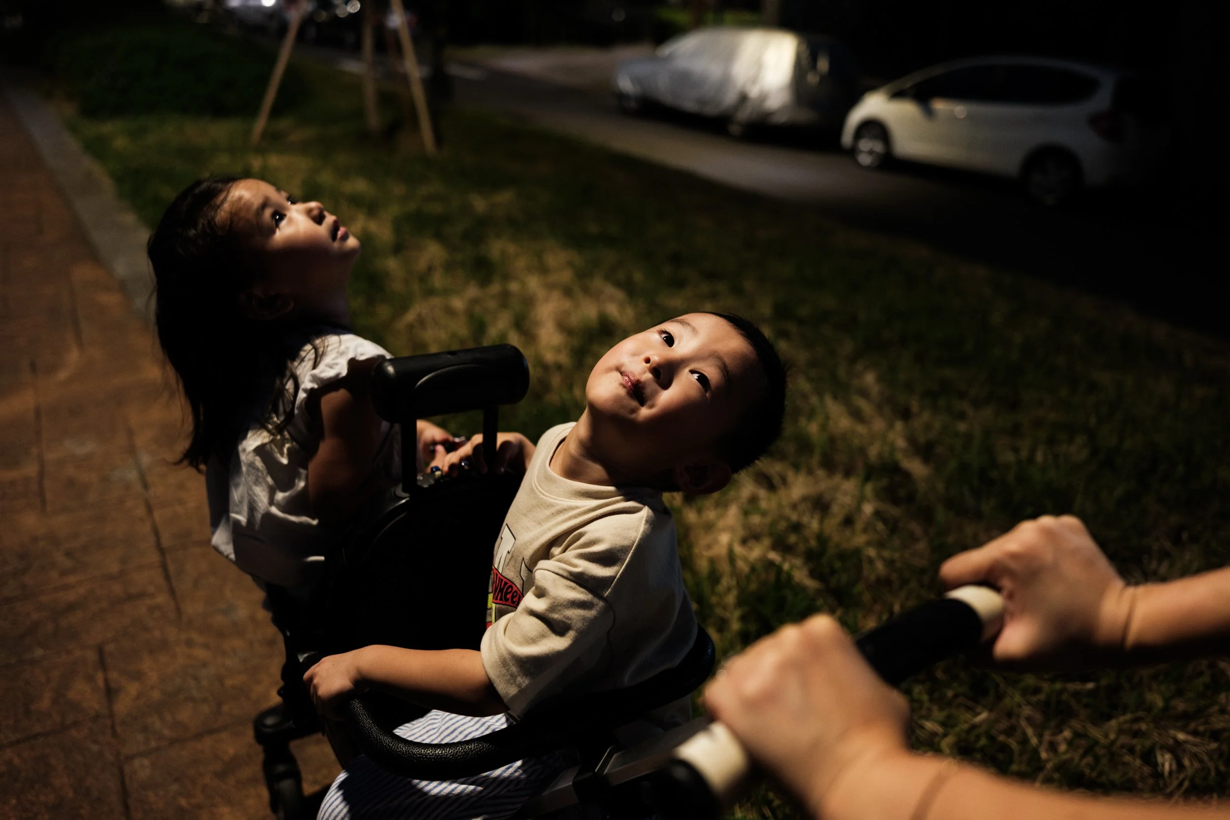 A color photo of a little boy and girl looking up towards a light source as they're pushed in a stroller along a Taipei riverside path.