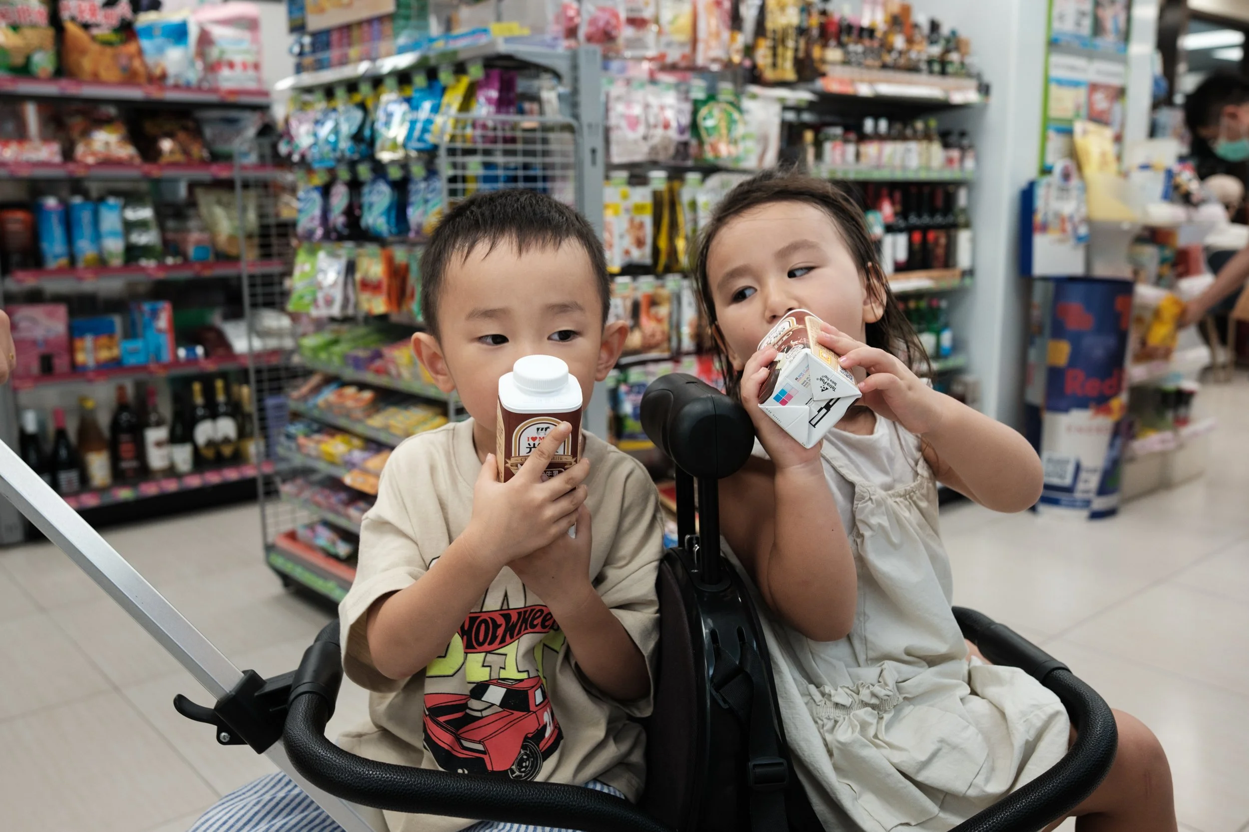 A color photo of a little boy and girl drinking milk in their stroller in a Taipei convenience store. Photographed by documentary family photographer Cahleen Hudson.