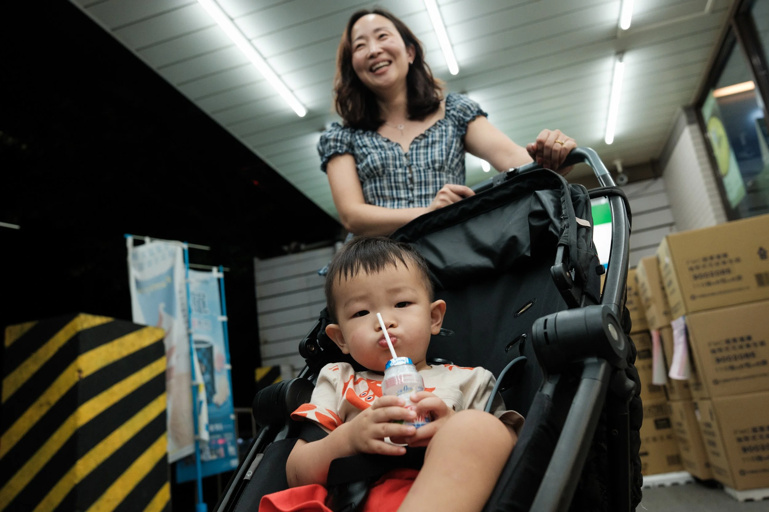 A color photo of a little boy sitting in his stroller and drinking milk from a straw outside a Taipei convenience store. His mom is standing aboce him with her hands on the stroller handles.