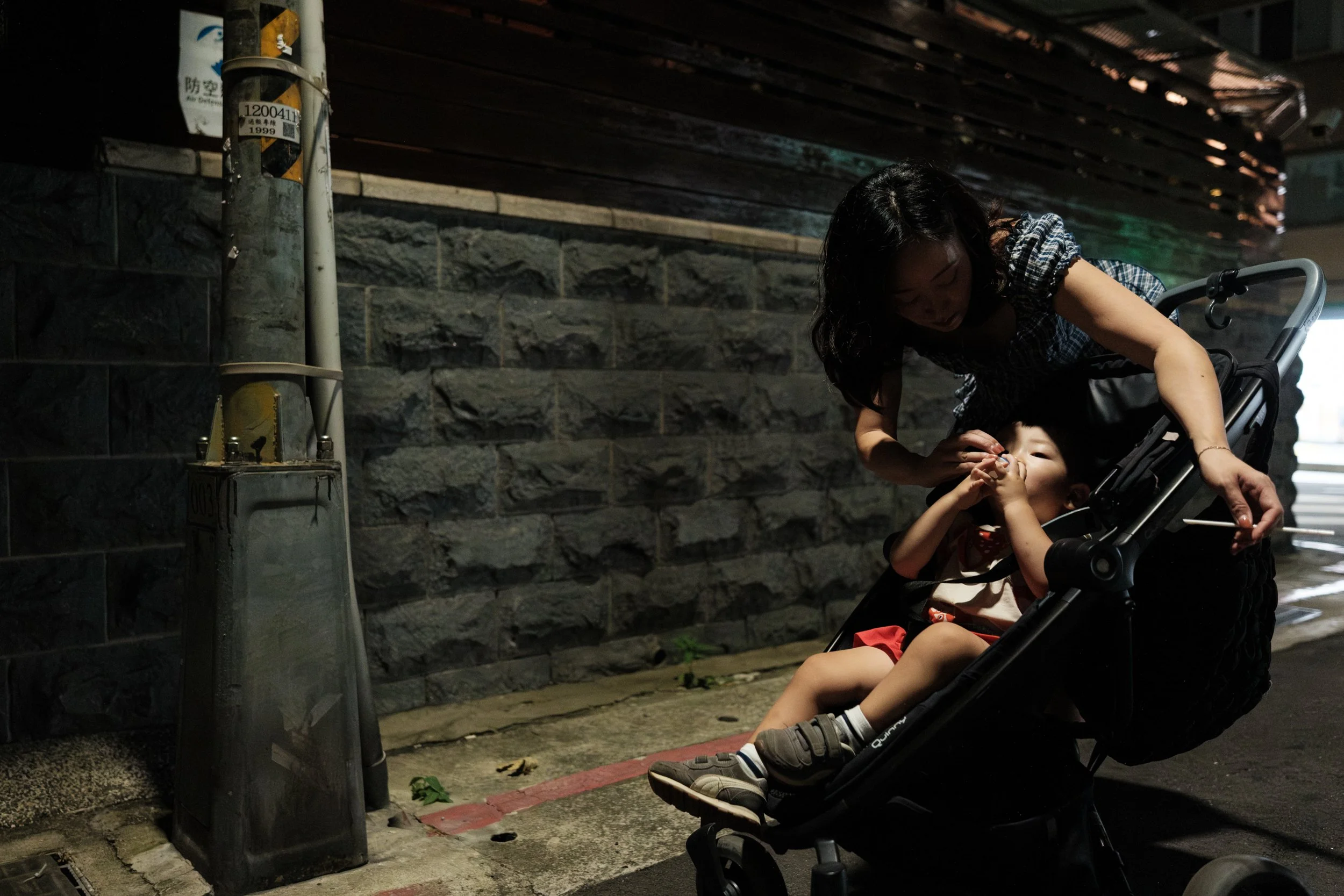 A color photo of a woman bending over a stroller and tending to her toddler son. They are standing underneath a street lamp on a Taipei street.