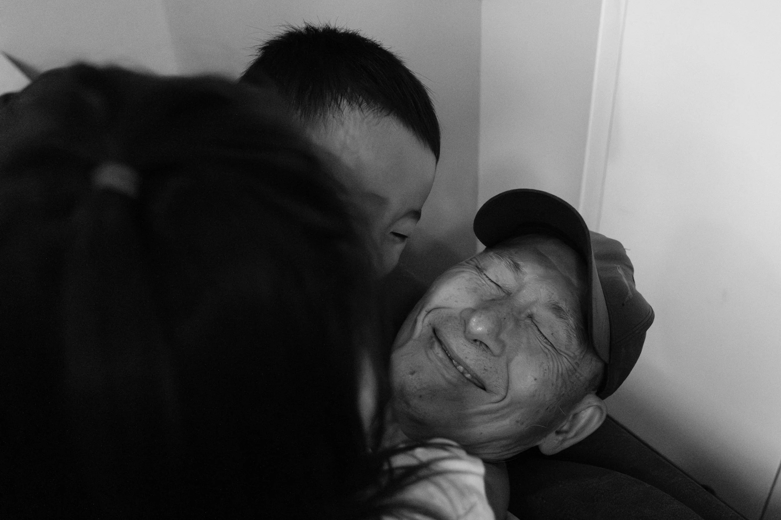A black and white photo of two kids jumping on their grandfather on a couch and hugging him. The photo focuses on the grandfather's face; he's smiling with his eyes closed. 