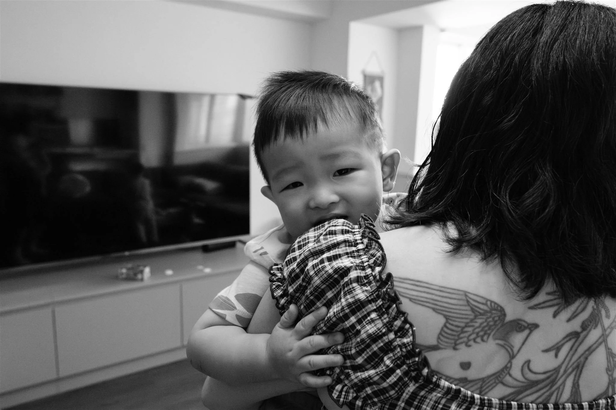 A black and white image of a little boy looking over his mom's choulder as she holds him. The mom has a big tattoo on her shoulder. Photographed by Taipei family photographer Cahleen Hudson. 