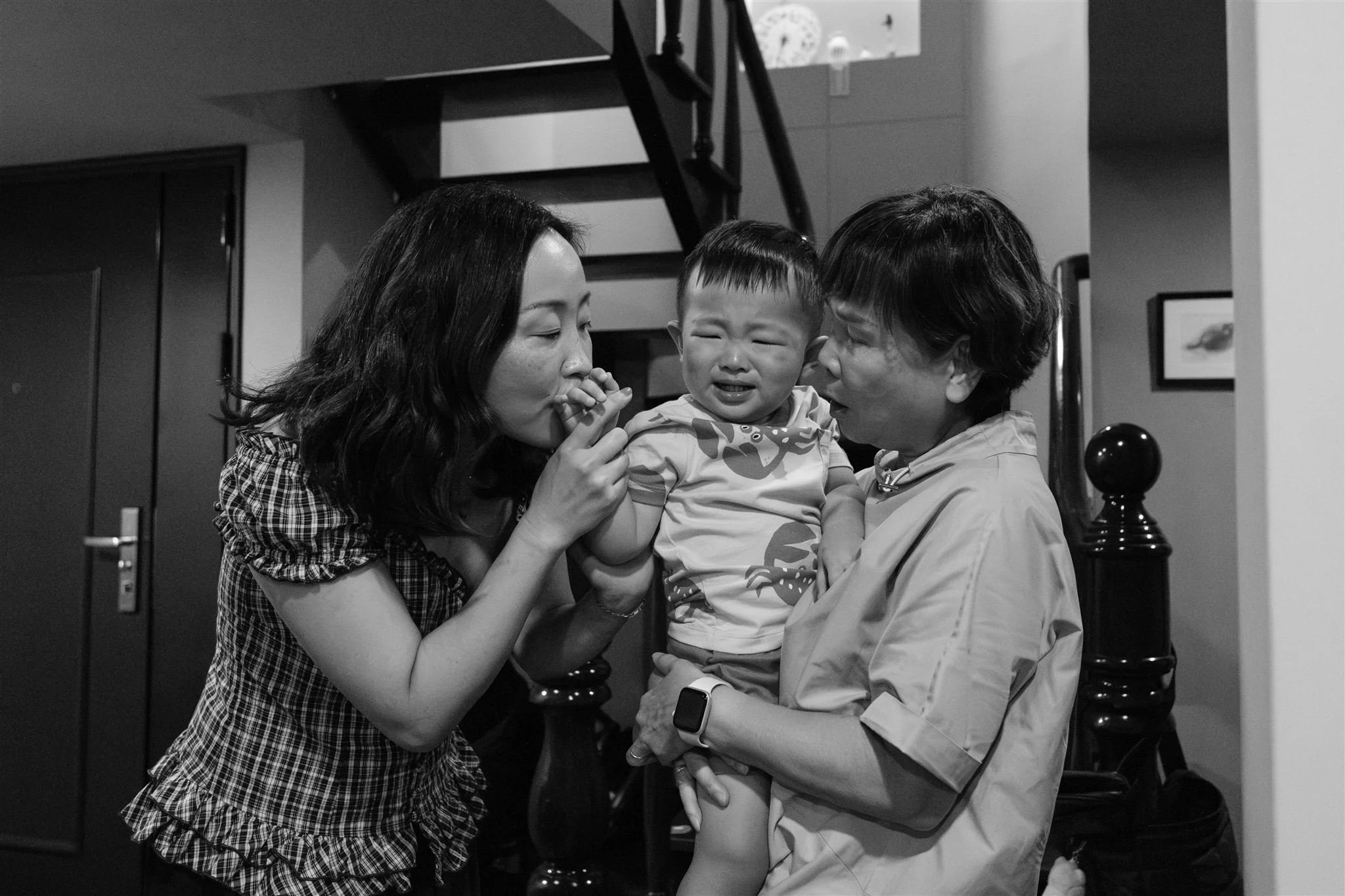 A black and white photo of a little boy who is crying and being held by his grandmother in the doorway of a Taipei apartment. The boy's mom is standing next to him and kissing his hand.