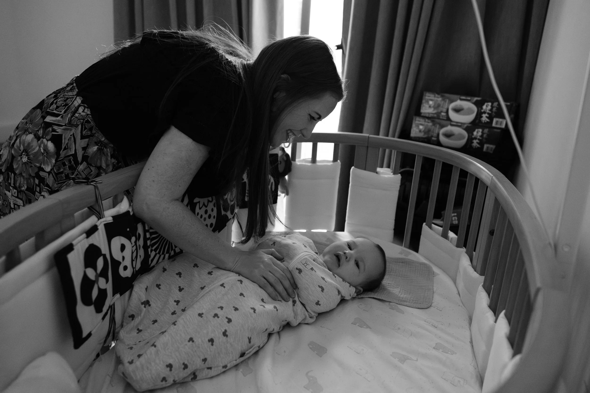 A woman leans over her baby in his crib and smiles at him as she picks him up. He just woke up from a nap. Photographed by Taipei photographer Cahleen Hudson.