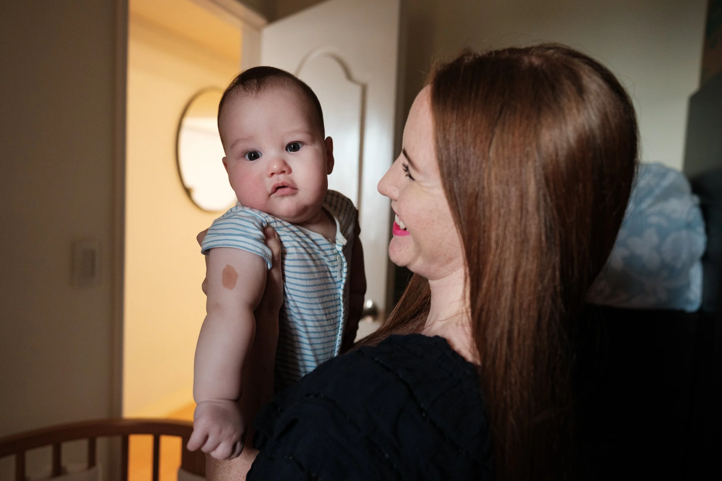 A color image of a woman holding up her baby in their bedroom in Taipei. Light from a window is shining on the baby's face.