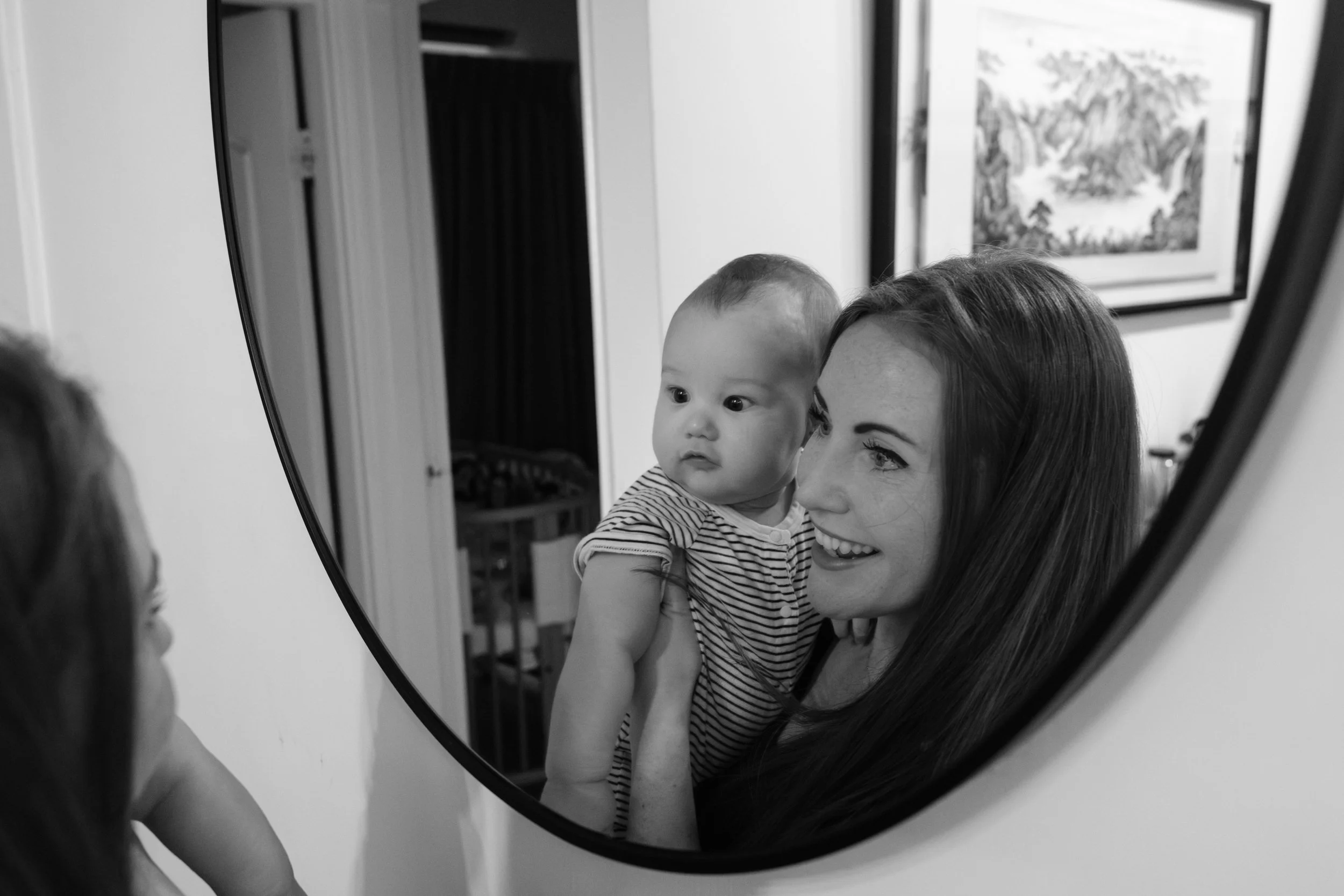 A black and white image of a woman holding her baby and looking in a mirror hanging on a wall in their Taipei apartment with him. The baby looks amused to see his reflection.