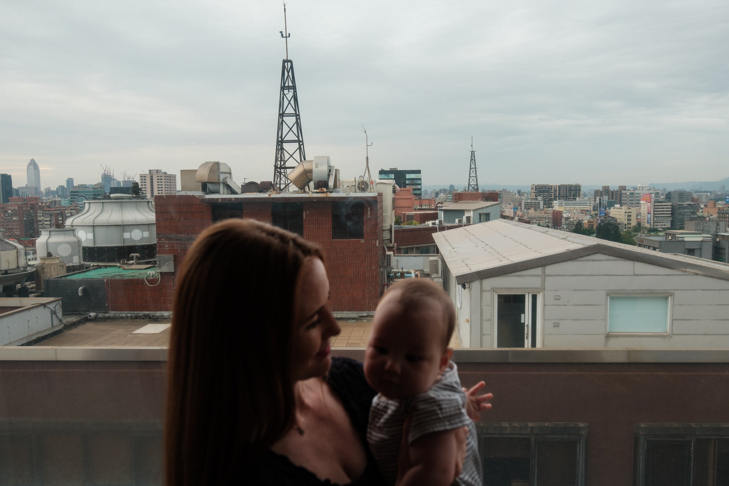 A color image of a woman holding her baby in front of a big window in their living room in Taipei. The focus is on the citny scene behind them. The woman is looking at her baby.