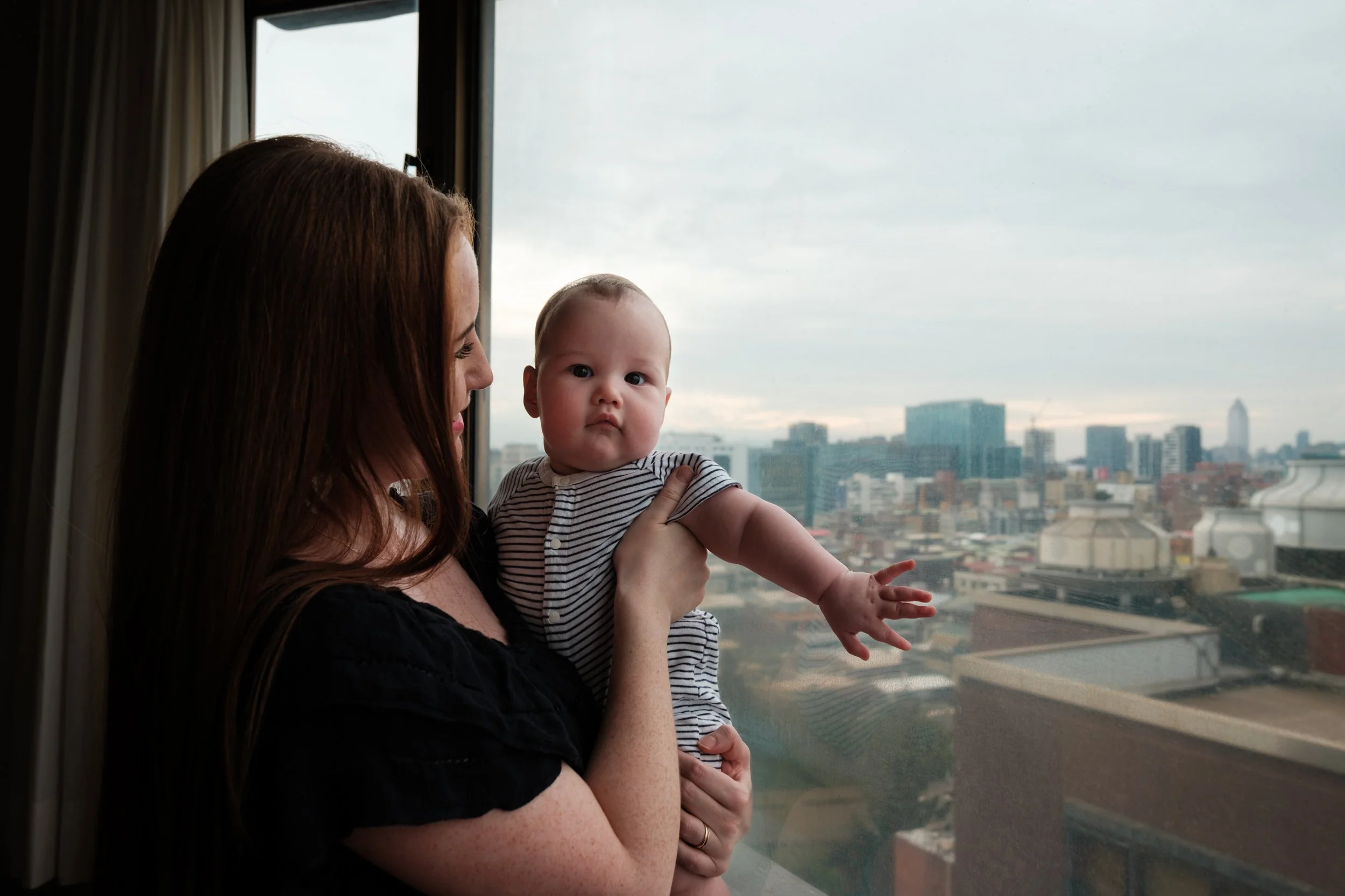 A color image of a woman holding her baby in front of a big window with a city view in their Taipei apartment. The woman is smiling at her baby and the baby is looking at the camera.