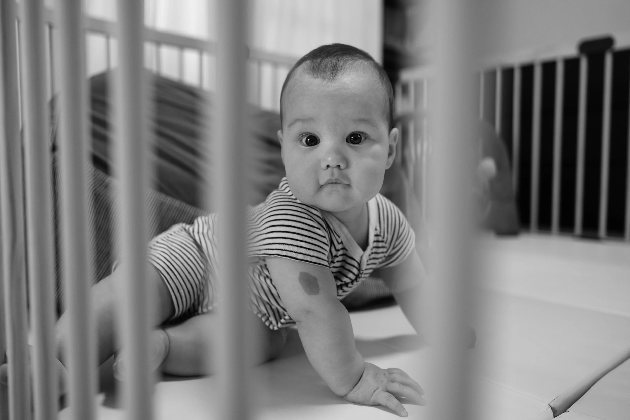 A black and white photo of a baby on all fours on his foam play mat. His face is framed between the bars of his play pen. Photographed by Taipei family photographer Cahleen Hudson.