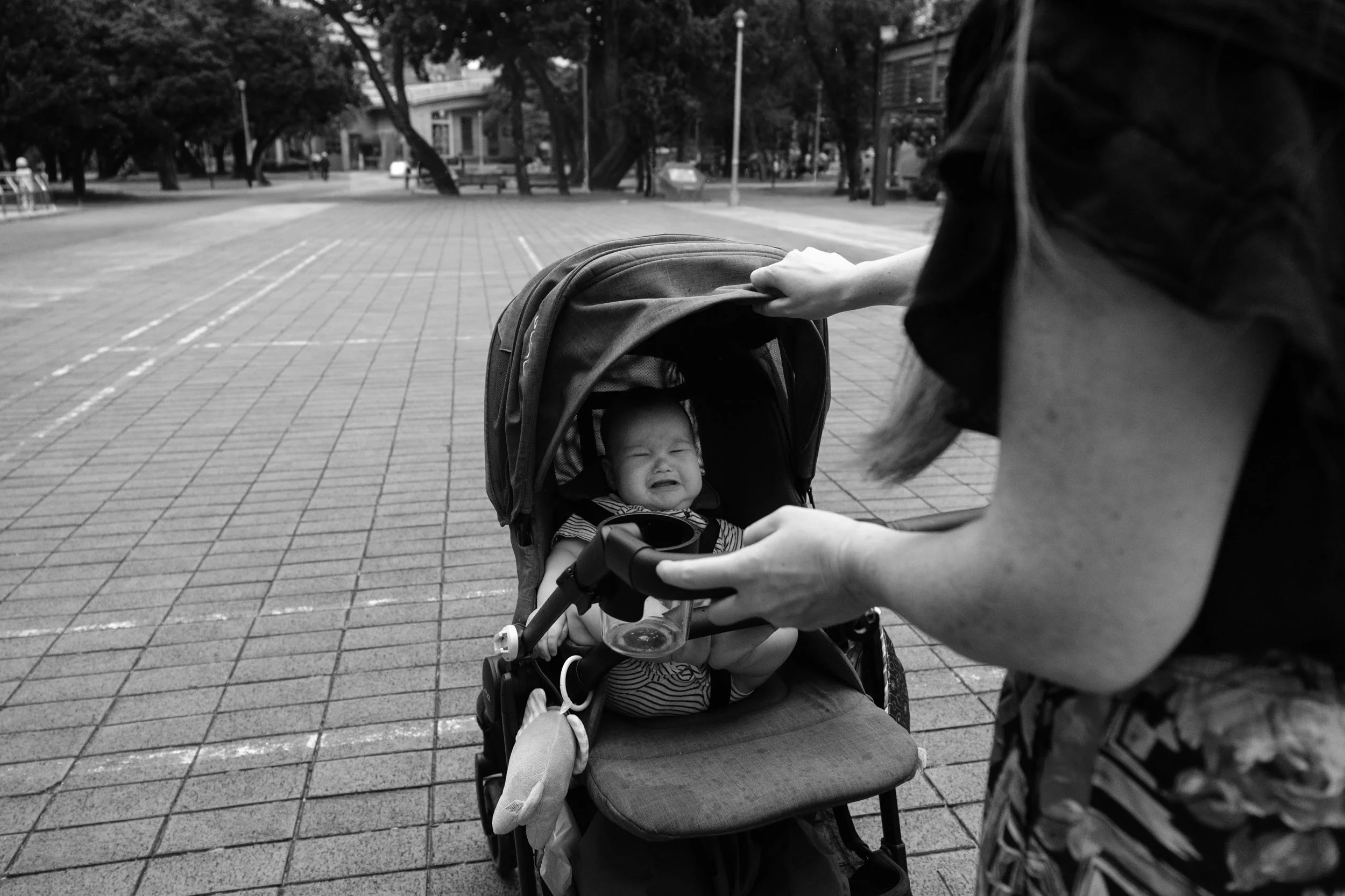 A black and white photo of a baby crying in his stroller as his mom adjusts the sunshade. Photographed in Rongxing Park in Taipei by Cahleen Hudson.