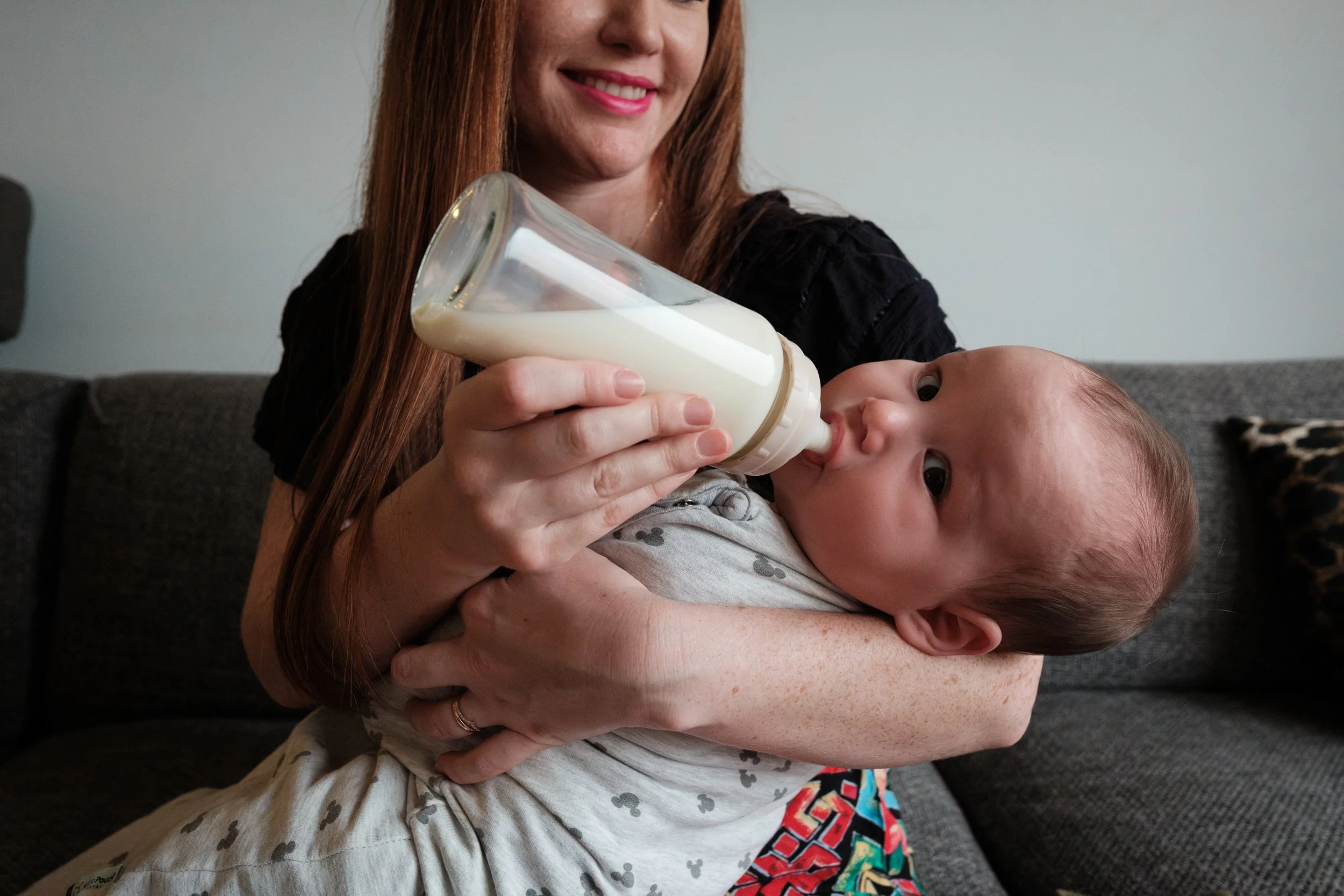 A color photo of a woman feeding her baby a bottle of milk on her couch in her living room in Taipei. The baby is getting distracted and turning away to look at the camera as the mom laughs. 