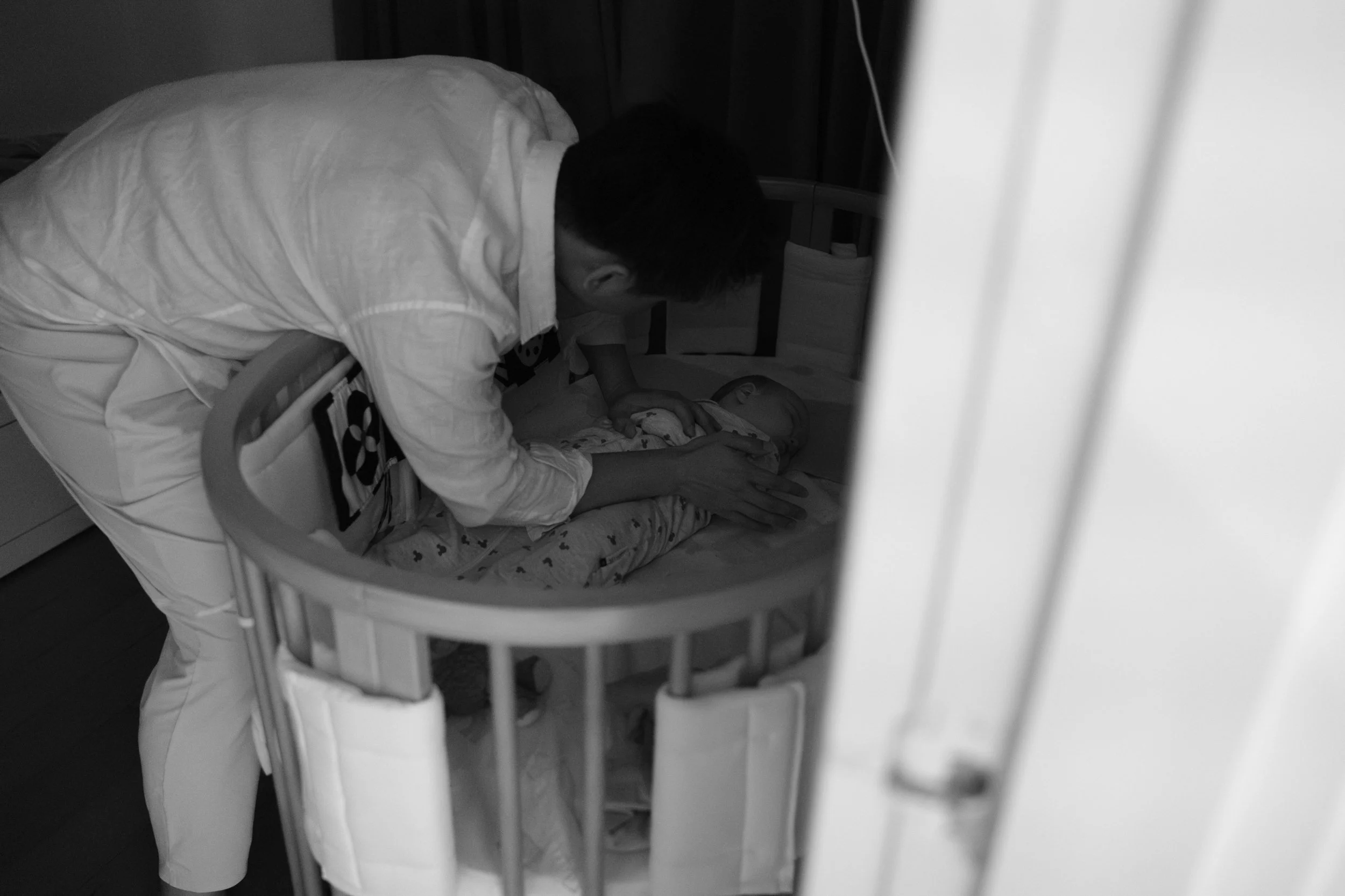 A black and white image of a dad carefully leaning over and placing his sleeping baby in a crib in a dark room. Photographed by Cahleen Hudson during a home family photoshoot in Taipei. 