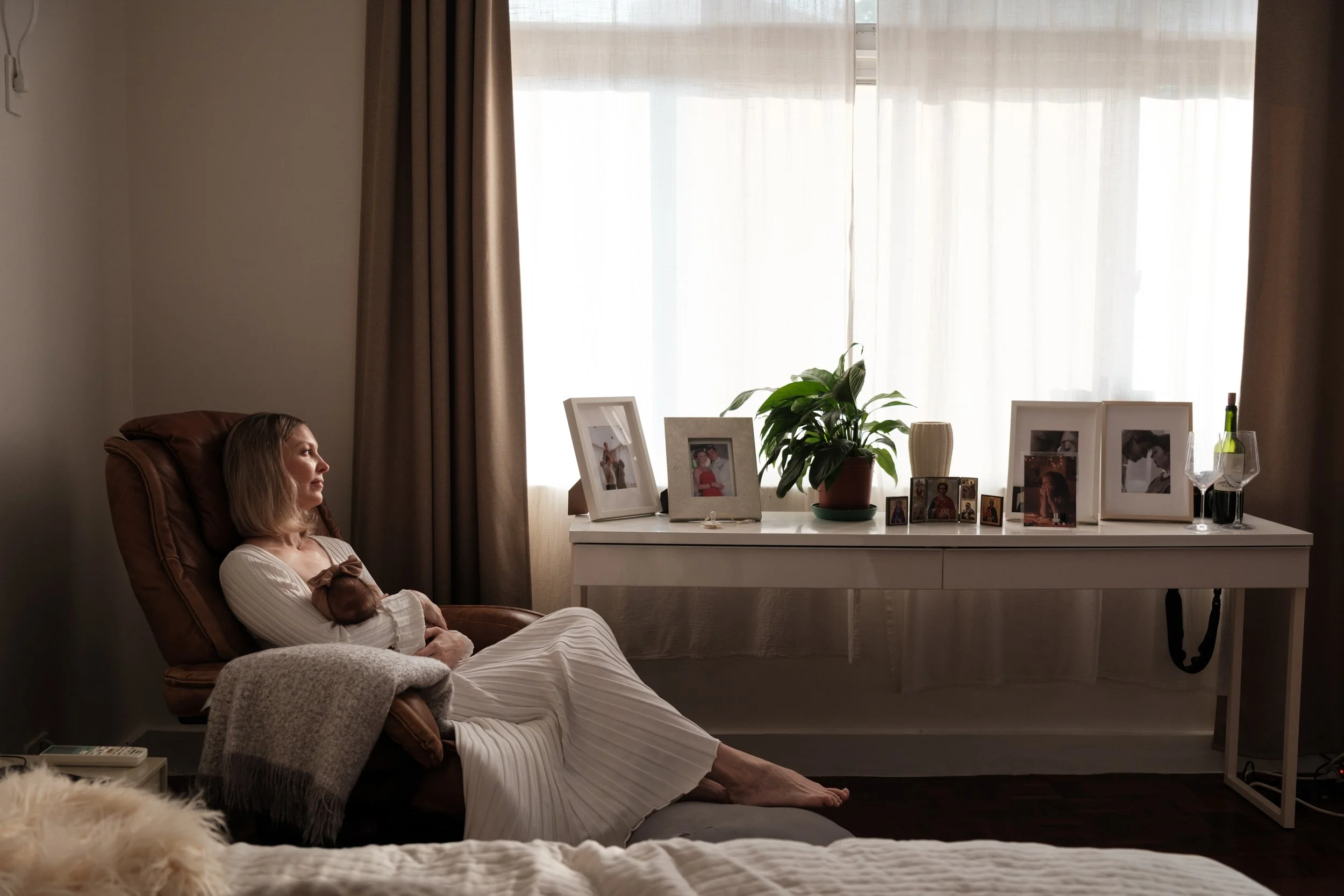 A mother gazes out of a window as she peacefully nurses her baby. She is next to a table with lots of framed photos.