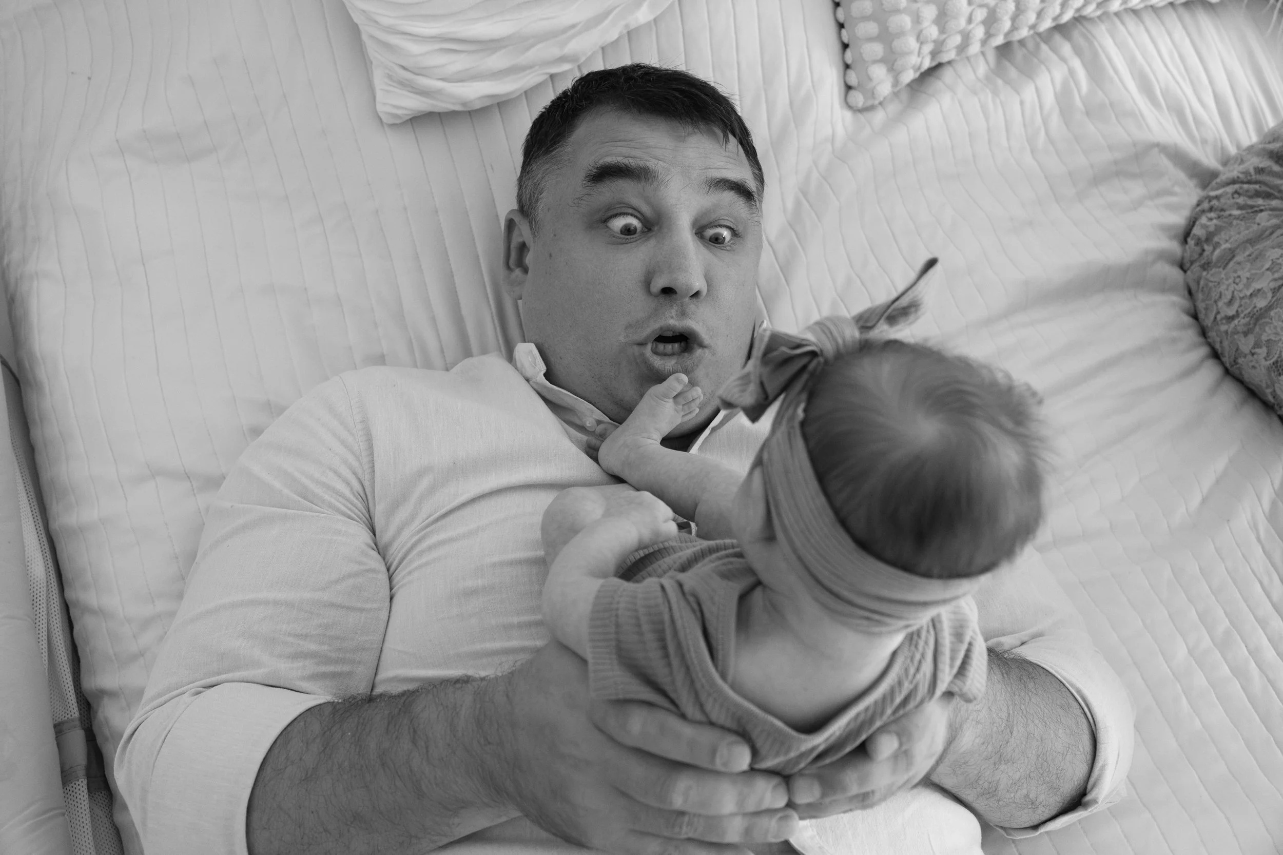  A father is lying on a bed and holding his newborn baby upright on his stomach as he makes a funny face. 