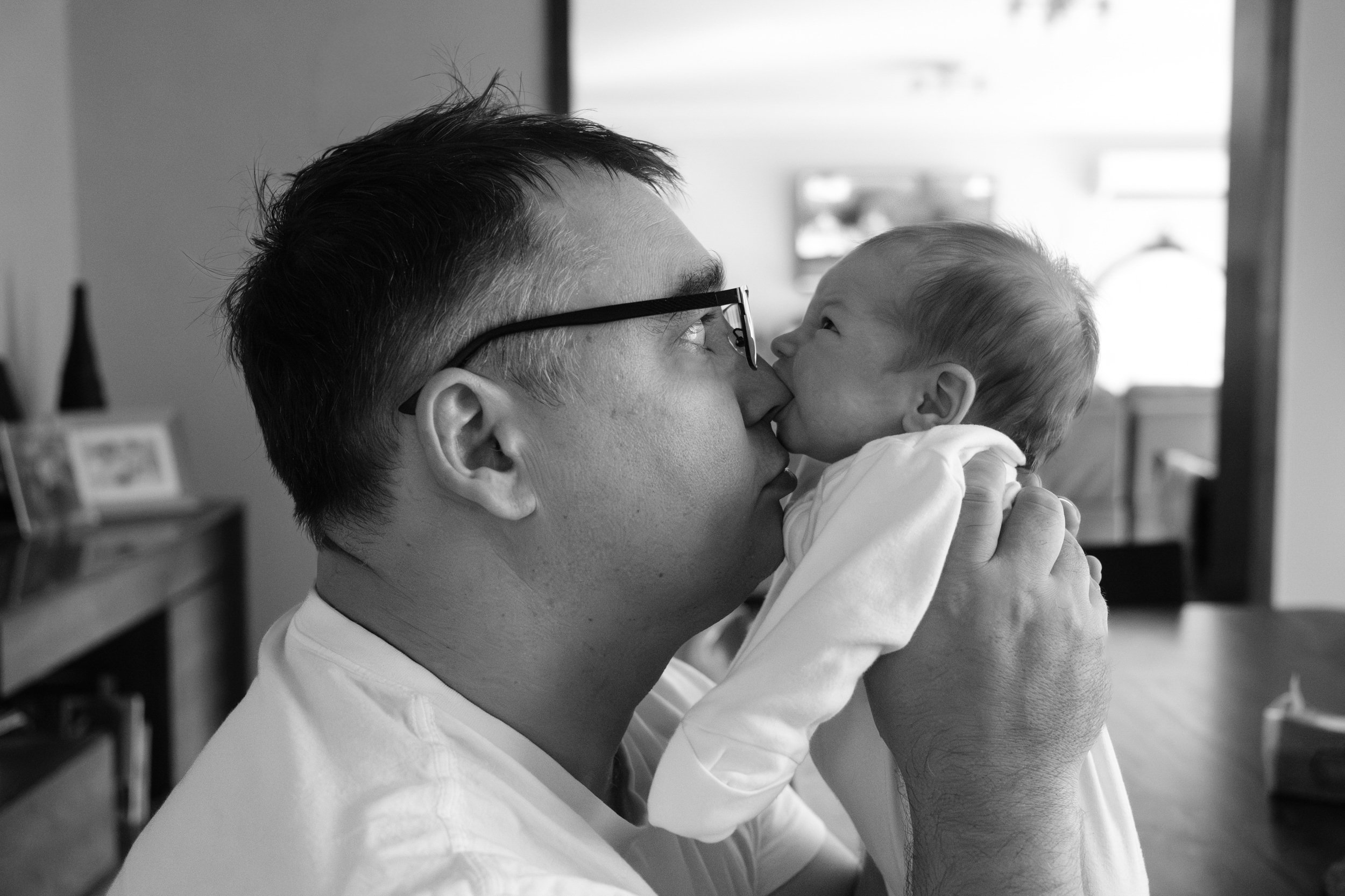 A black and white photo of a man holding his newborn baby daughter up to his nose. He is making a funny face as she tries to suckle his nose. 