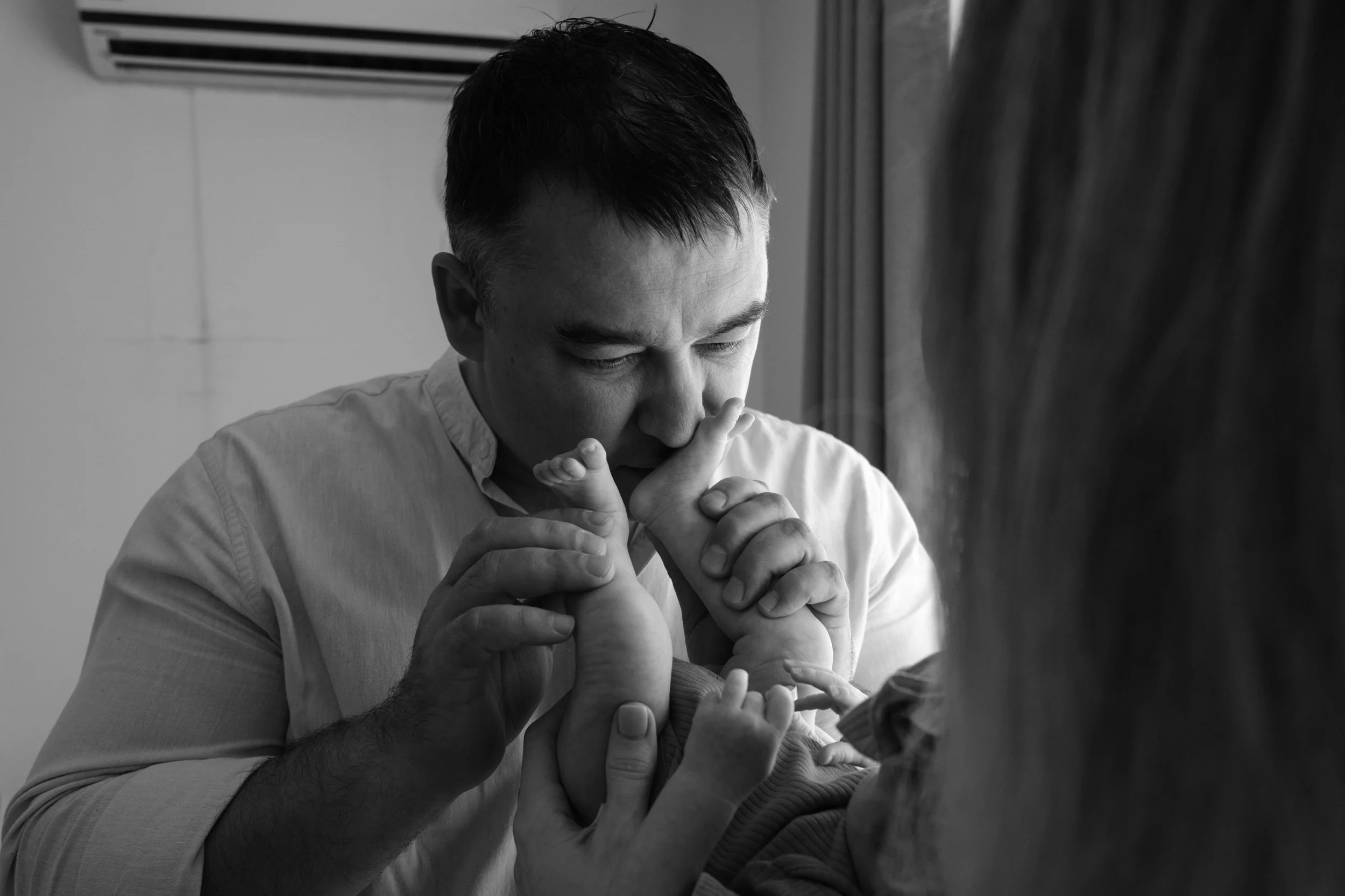 A mother is holding up her newborn baby as the father grab's the baby's legs and kisses the tiny feet next to a window. 