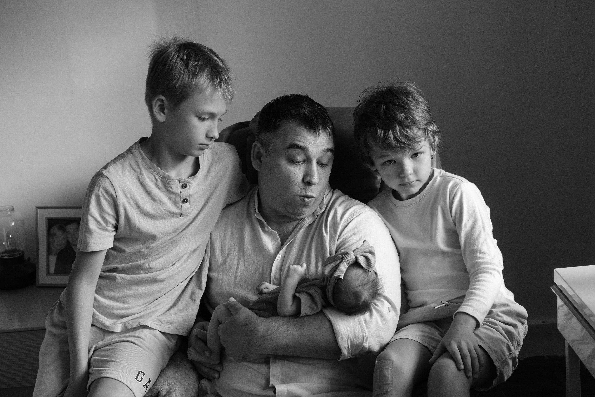 A father is holding his newborn baby in a rocking chair and making a funny face at her. The baby's older brothers are sitting on either side of the father on the arms of the chair.