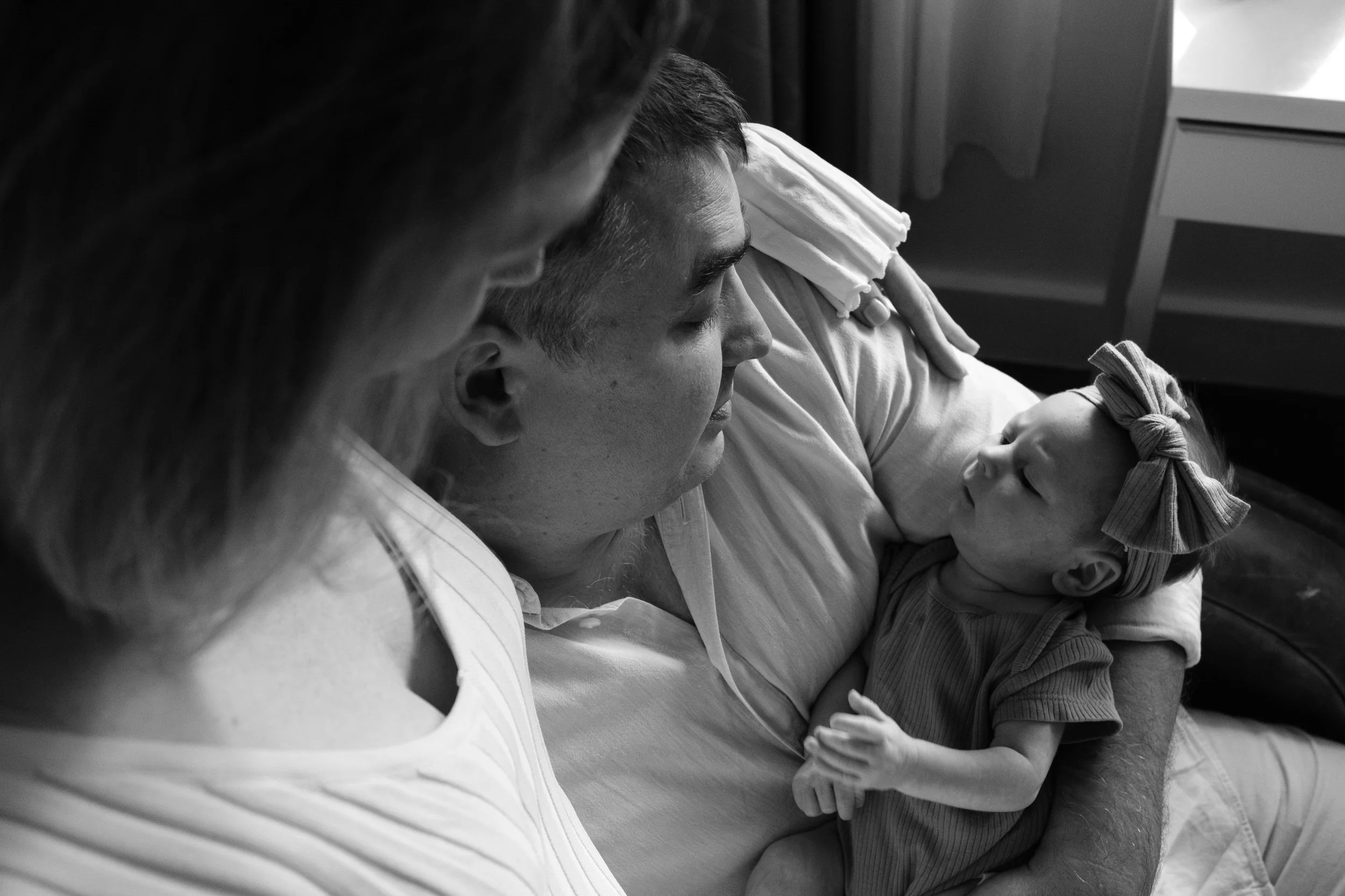 A father is holding his newborn baby in a rocking chair by a window. His wife is sitting on the arm of the chair and they are both looking down at their baby daughter.