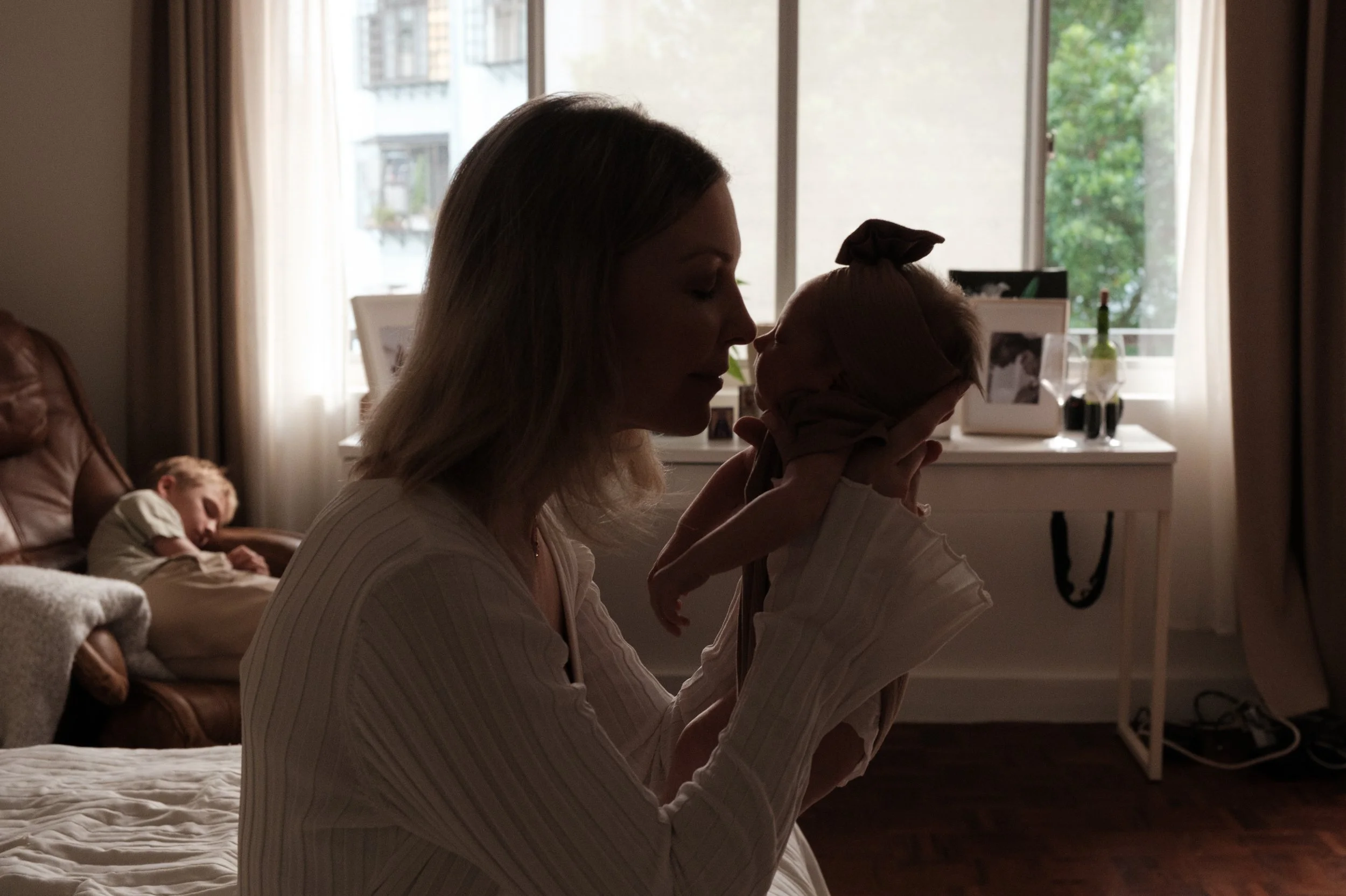 A mother is holding up her tiny baby daughter and touching noses with her. A big window is behind them.