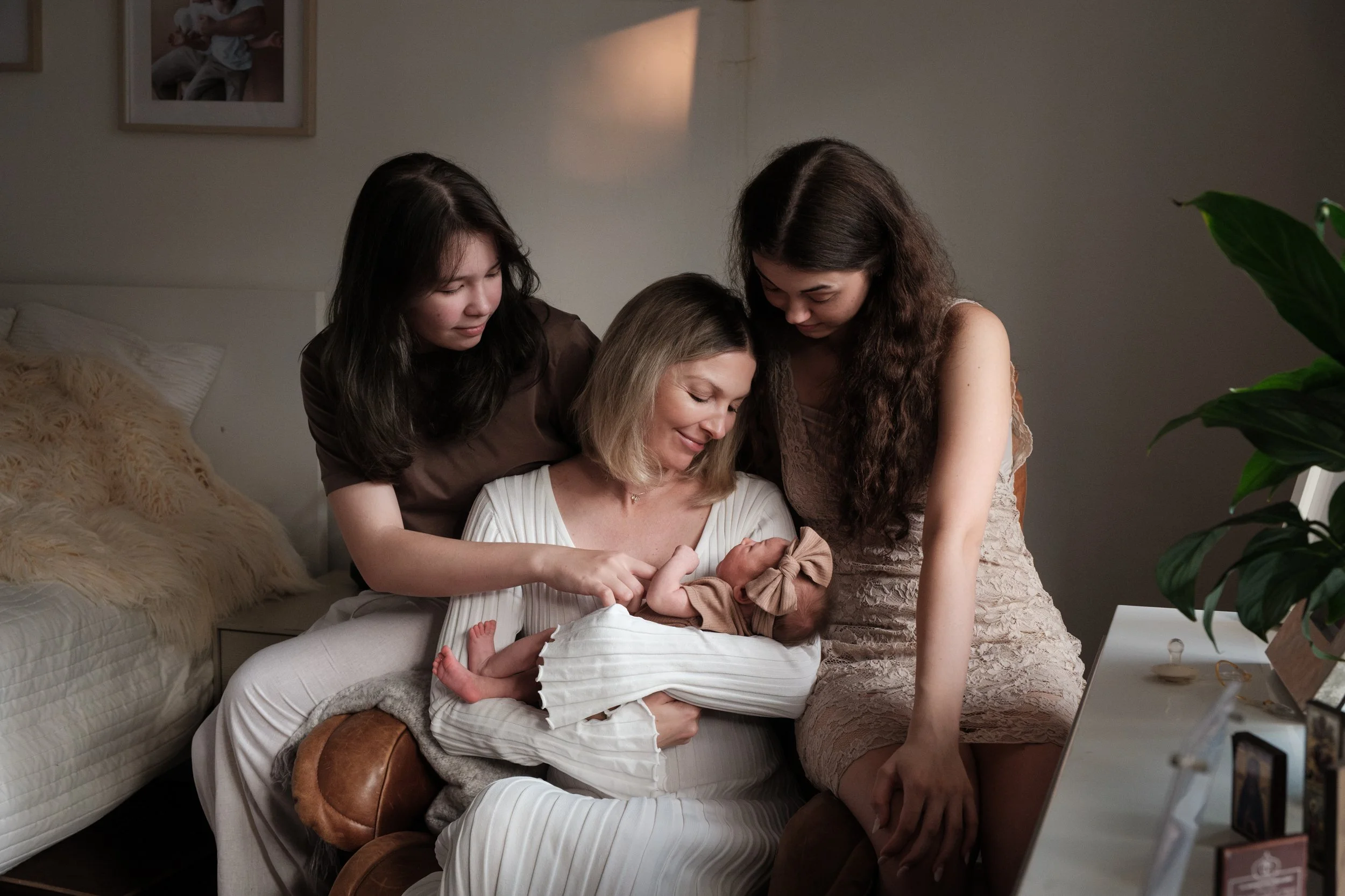 A mother in a white dress is holding her baby in a rocking chair and gazing down at her with a smile on her face. An older sister is sitting on each arm of the rocking chair. 