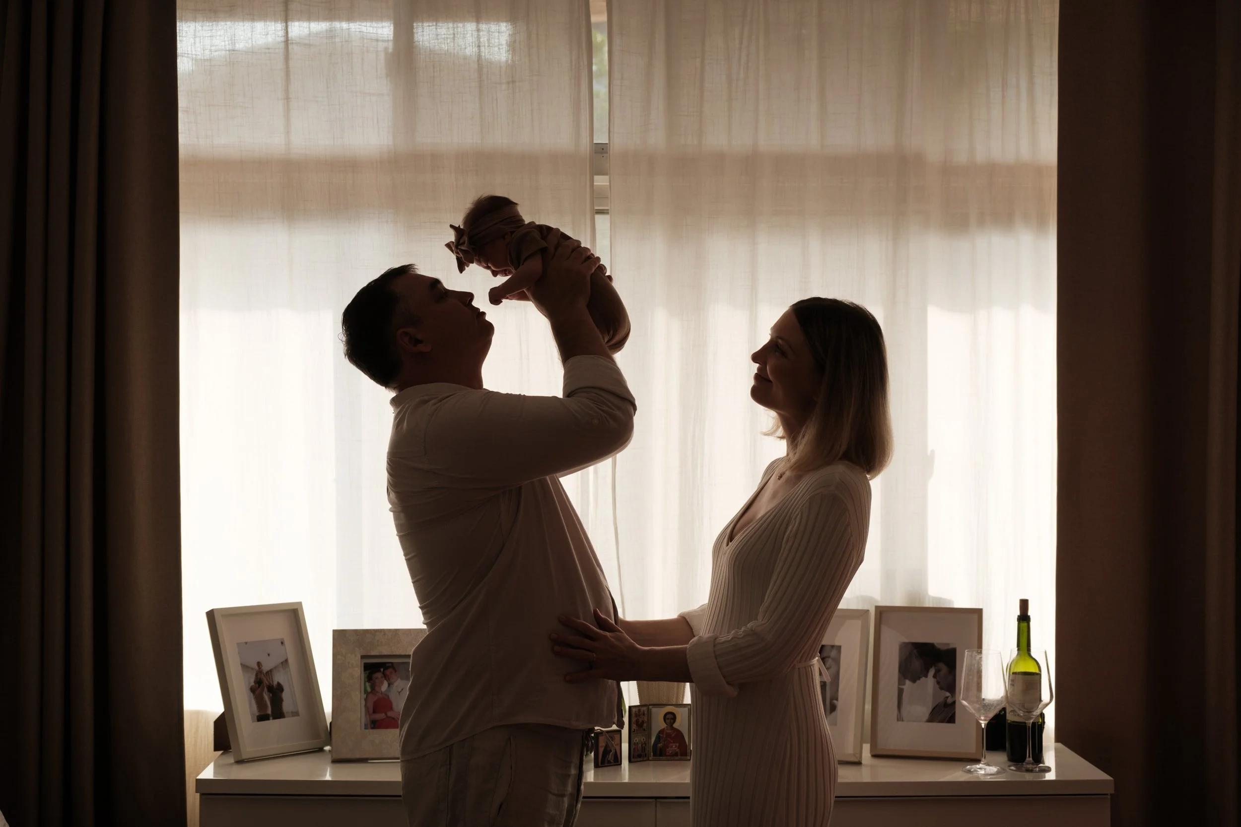 A father and mother are standing in front of a big window in their bedroom in Taipei and holding up their newborn baby. The father and baby are about to touch noses.