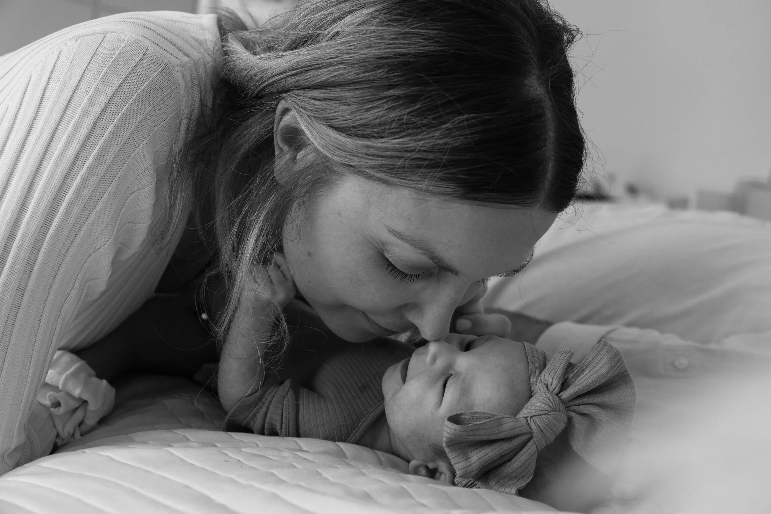 A newborn baby girl is lying on a bed and her mother is leaning over her and nuzzling her nose with her nose.