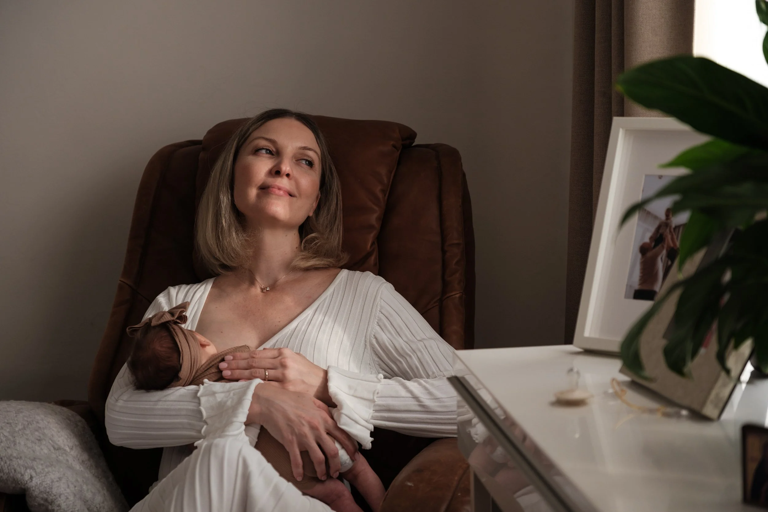 A mother smiles as she peacefully nurses her newborn baby girl by a window. The light from the window is washing over her and her baby.