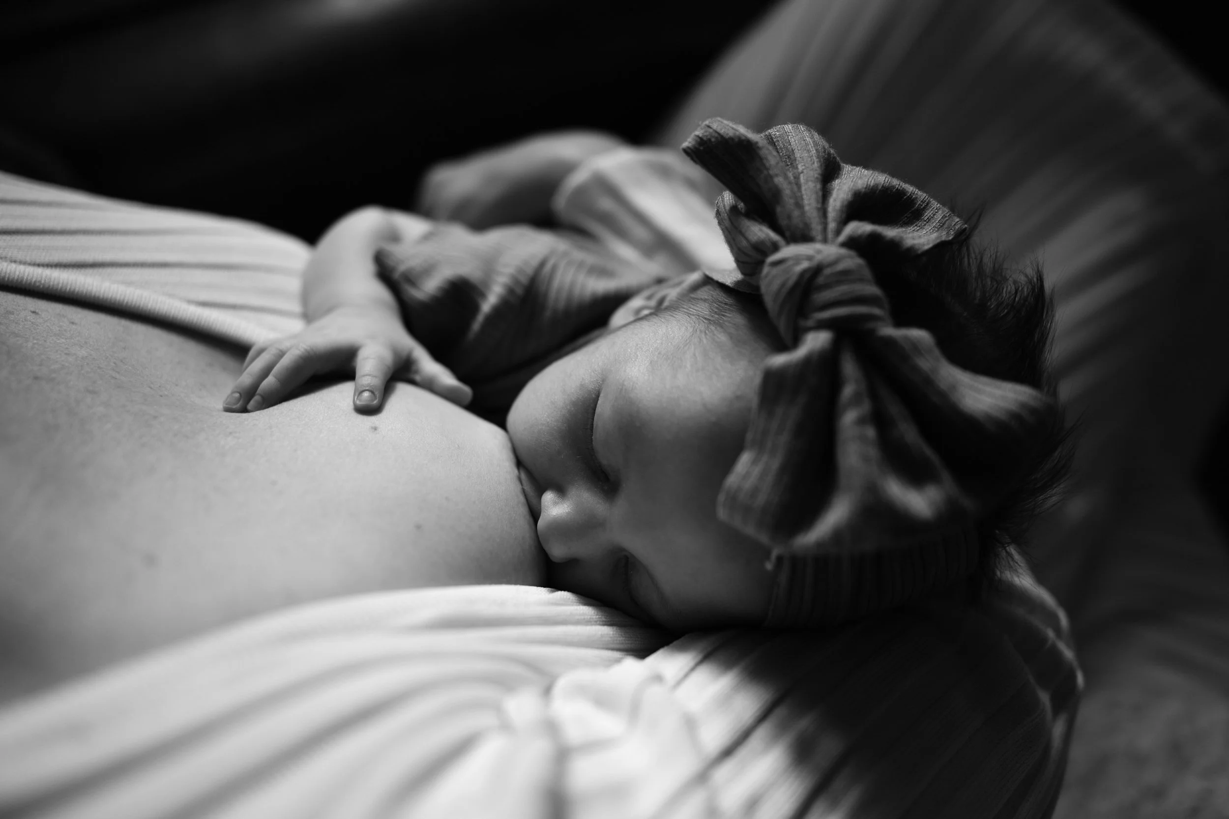 A newborn baby girl with a big bow on her headband is clutching the top of her mother's breast as she nurses.
