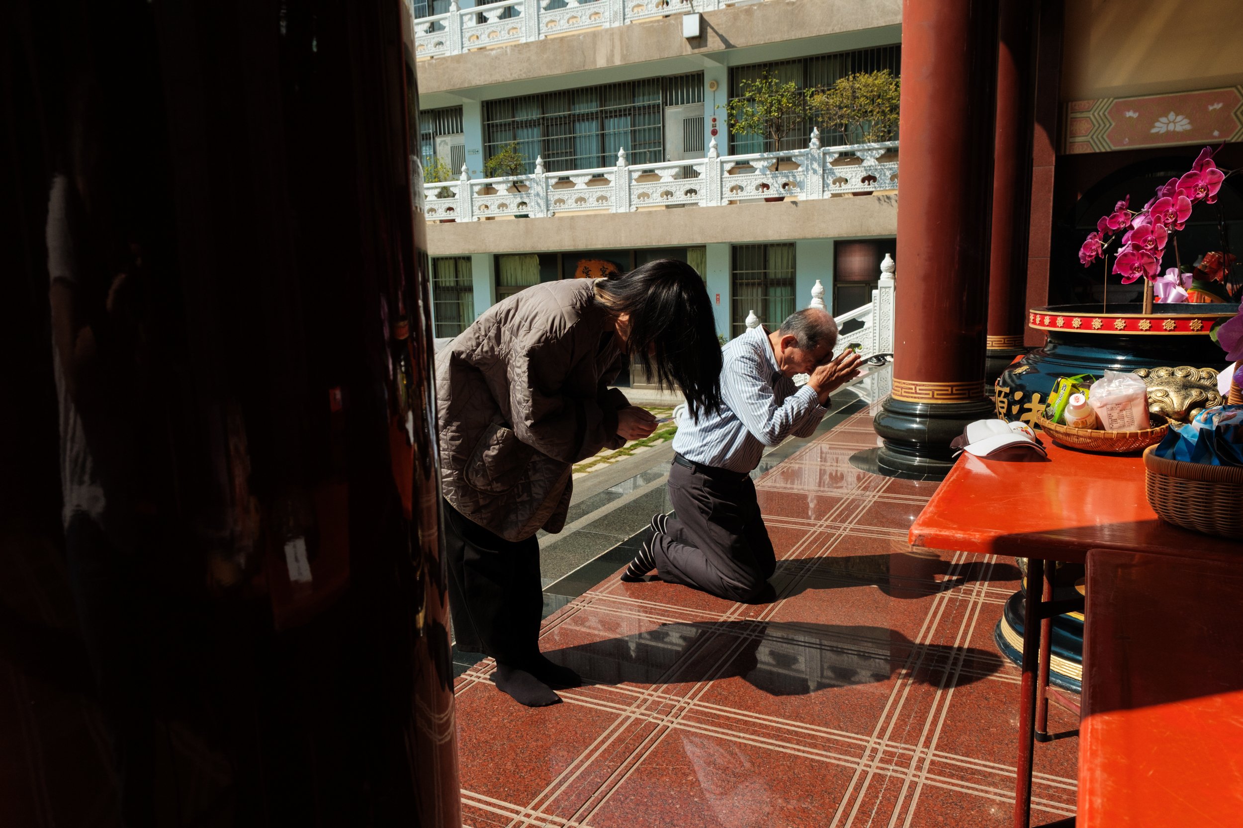 A woman is bowing to an alter with incense at a temple in Tainan. A man is kneeling on the ground next to her. A candid moment photographed during a travel family photography session.