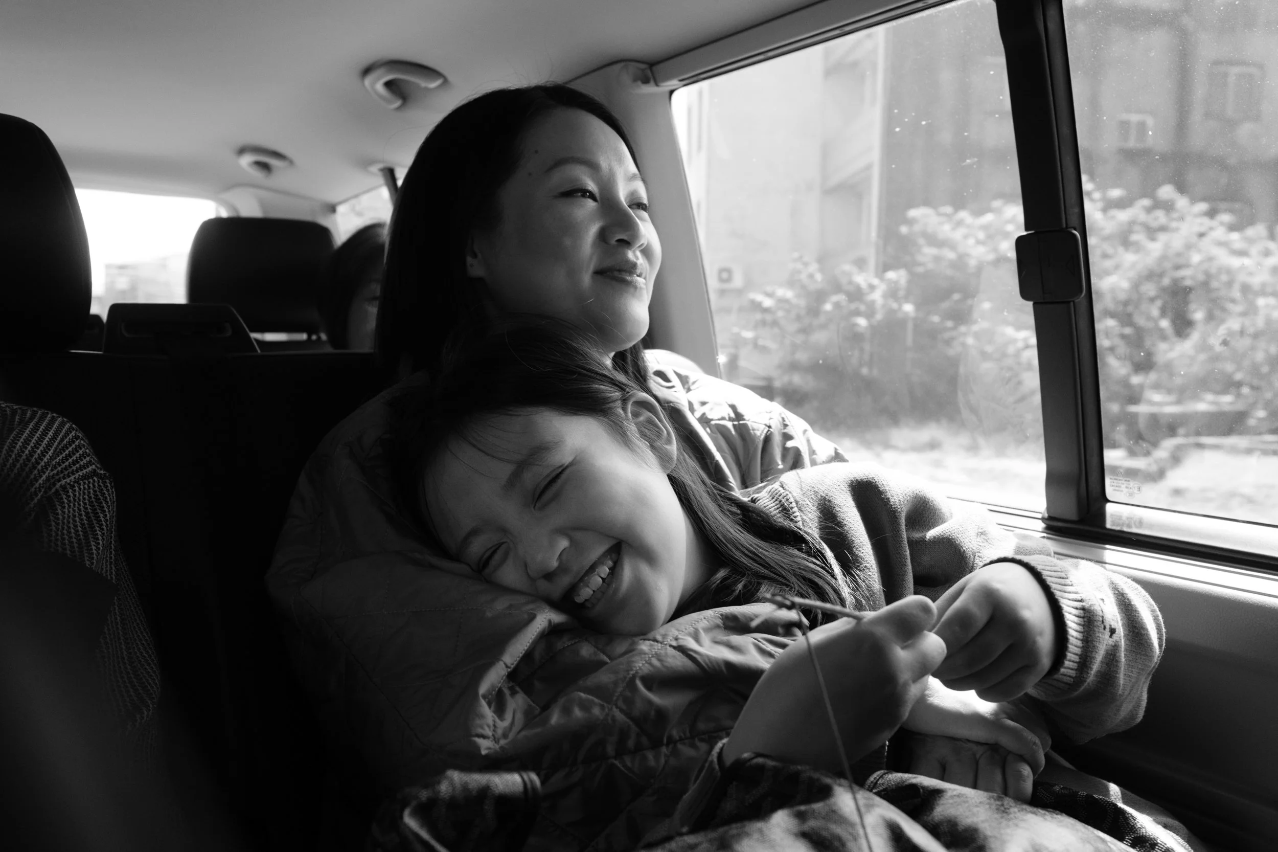 A woman is holding her little girl in the backseet of a car and smiling. The little girl is leaning against her mother and laughing. A natural family moment photographed in Tainan, Taiwan.