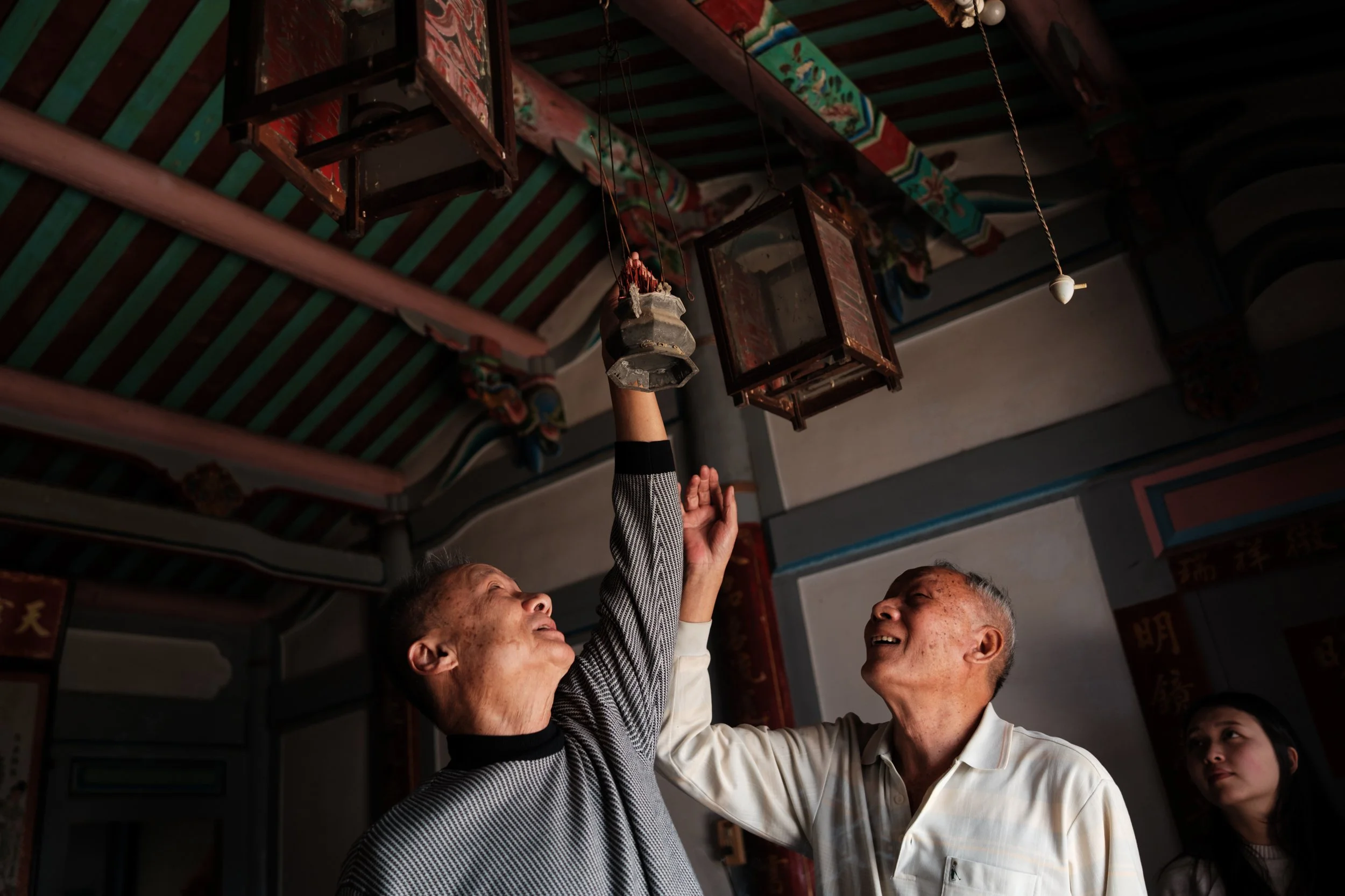 Two older men are holding incence and reaching up to place it in an incense holder hanging from the roof. 