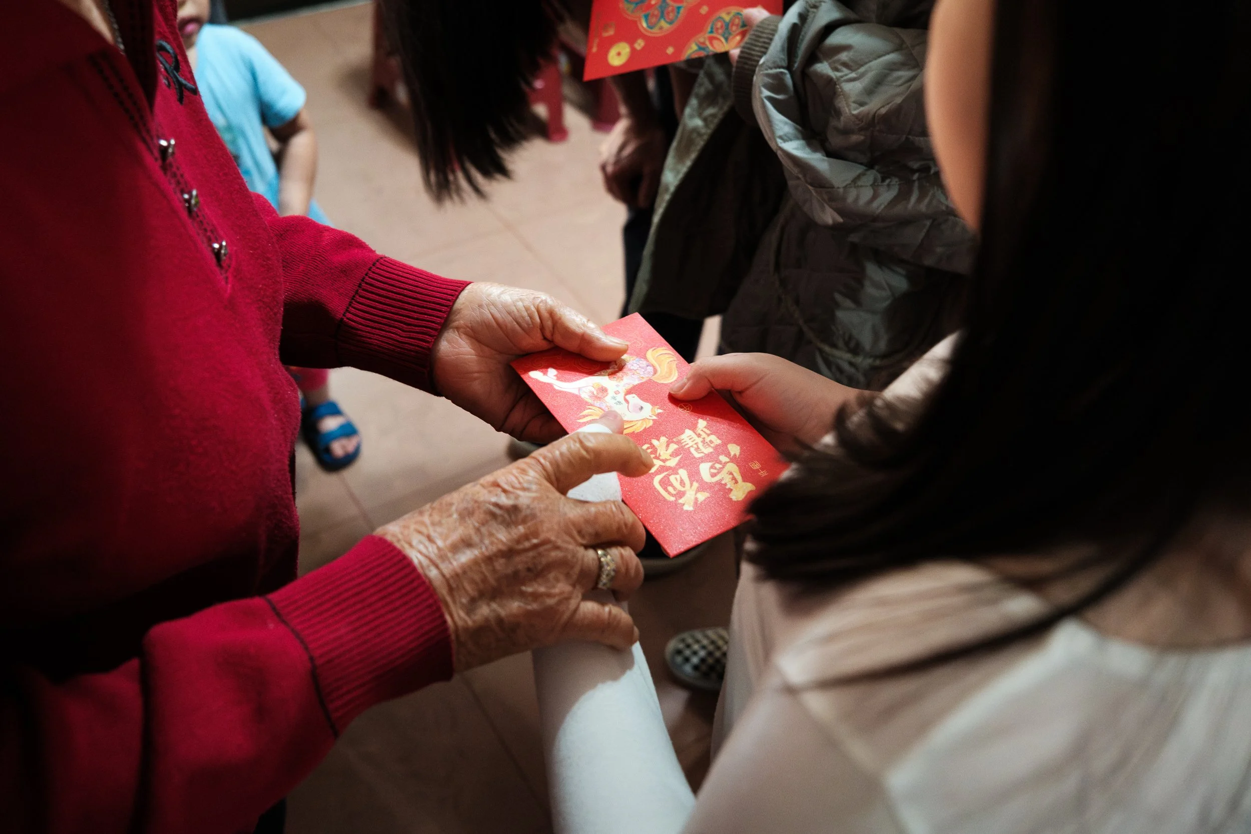 An old woman's hands are giving a red envelope to a young girl. A sweet moment where one of the old woman's hands are touching one of the young girl's hands.
