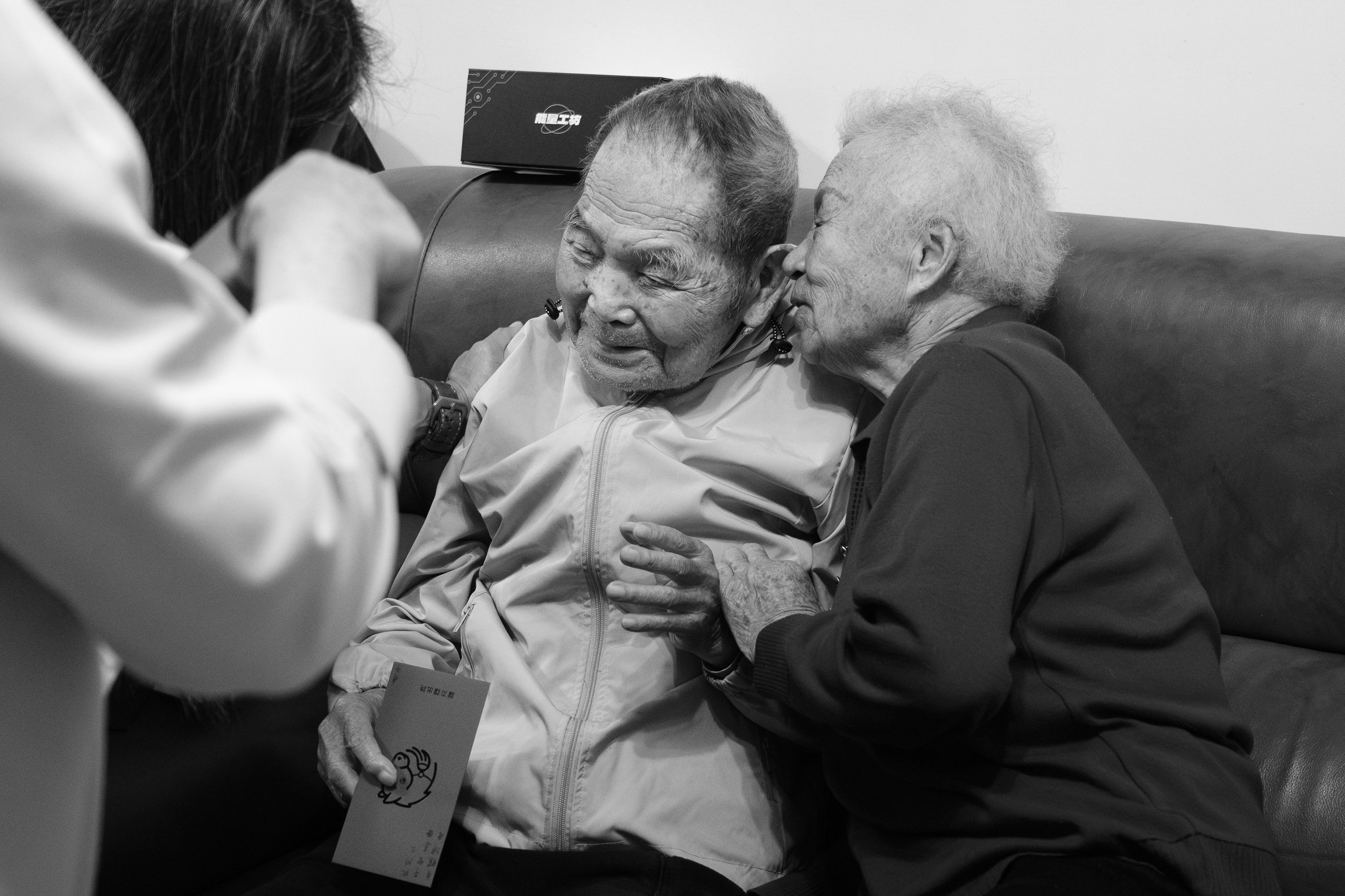 An old woman is sitting on a couch with an old man and whispering in his ear. A tender moment photographed during a day in the life session with Taipei family photographer Cahleen Hudson.